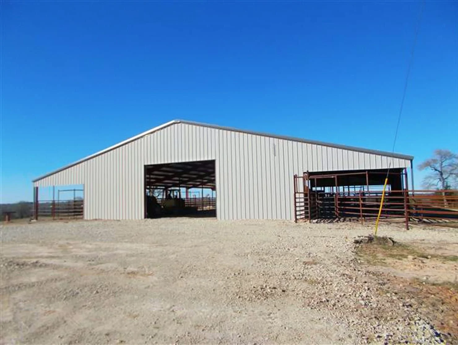 A large metal barn on a dirt lot with a clear blue sky overhead and some fencing around the structure.
