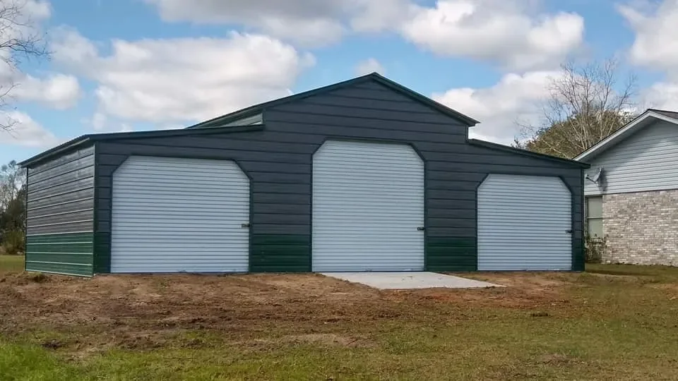 A large metal storage building with three garage doors, situated on a grassy area with a concrete pathway at the front, under a partly cloudy sky.