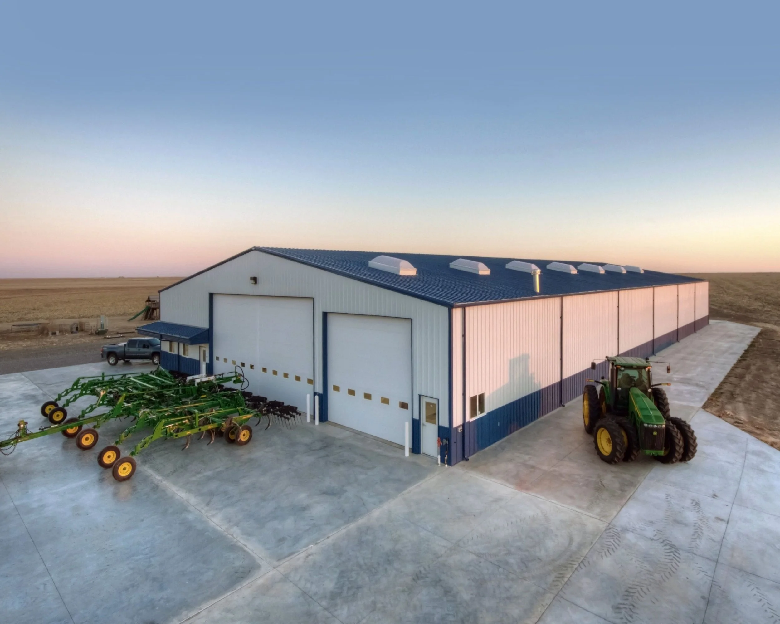 A large barn on a farm with a green tractor parked outside and farming equipment on the concrete pad, set against an open field at sunrise or sunset.