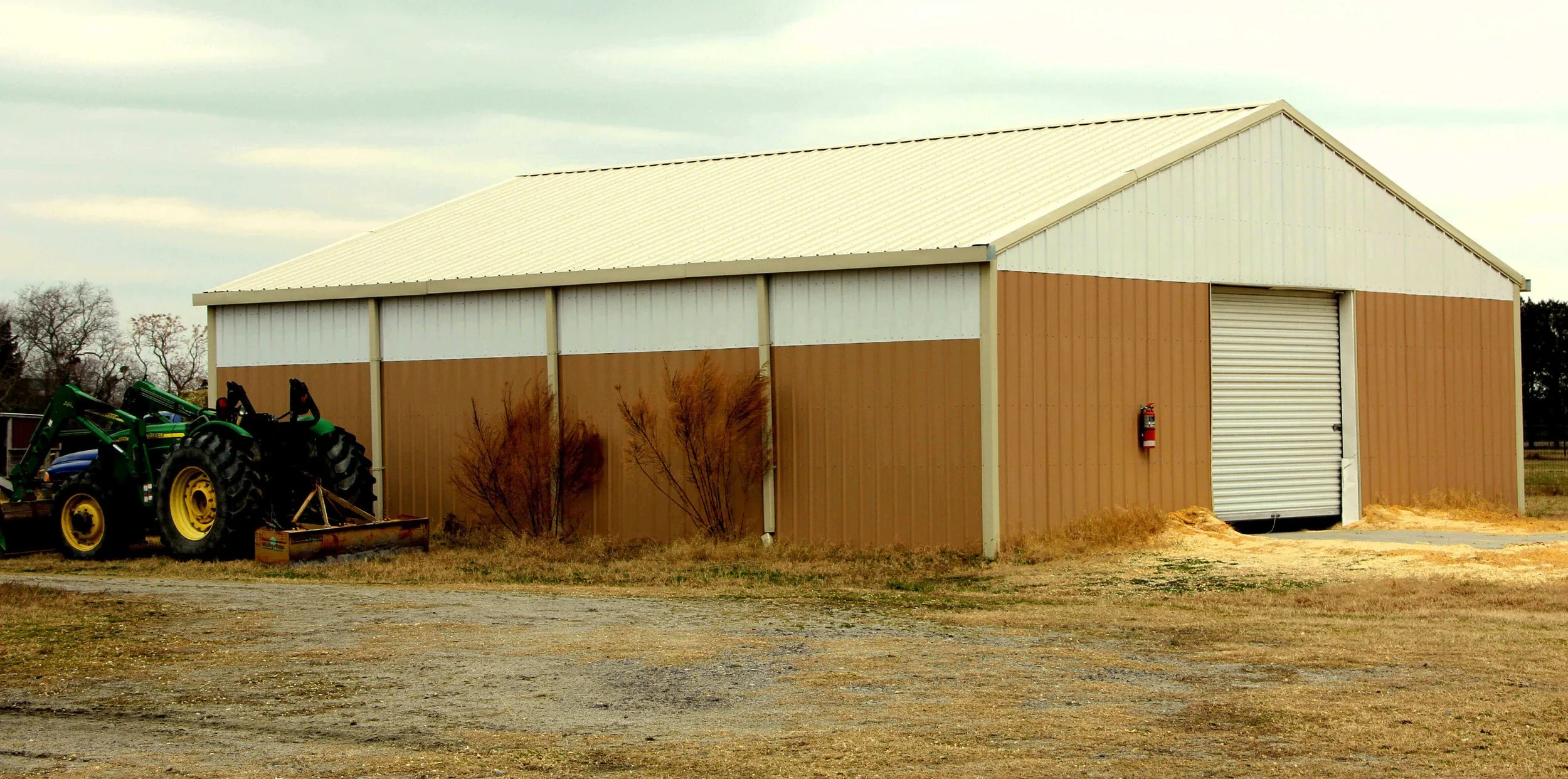 A large brown and white metal storage building with a roll-up door, a fire extinguisher mounted on the exterior wall, and a green tractor with a front loader attachment parked nearby.