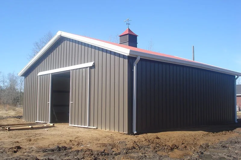A metal barn with a red gable roof, a cupola with a weather vane on top, and an open sliding door, situated on a farm or rural land under a clear blue sky.