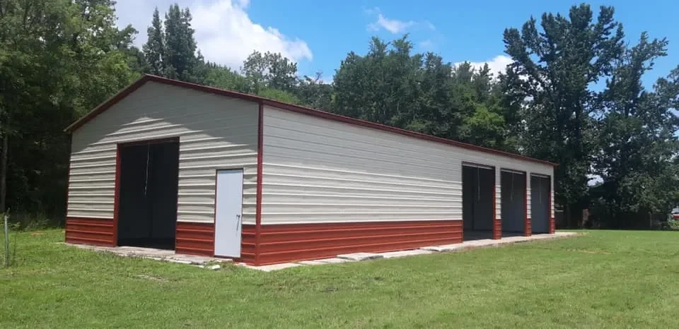 A metal storage building with four garage doors, situated on a grassy lawn with trees in the background under a partly cloudy sky.