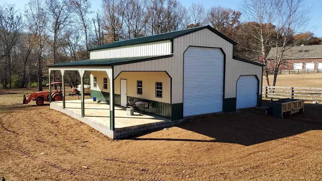 A large metal barn with a green roof and trim, featuring two garage doors and a small porch area with tools and equipment. There is a tractor parked nearby, and the ground around is bare dirt, with a wooded area in the background.