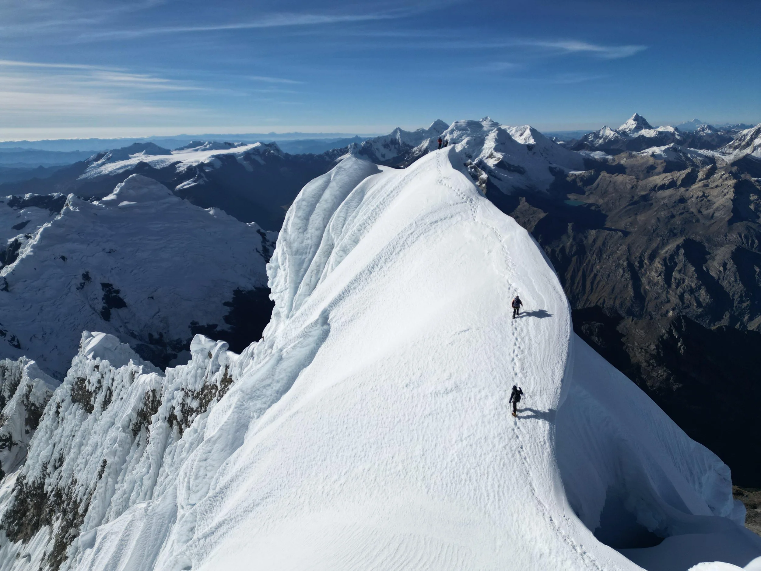 Three climbers are ascending a snow-covered mountain ridge in a high-altitude environment, surrounded by distant mountain ranges under a clear blue sky.