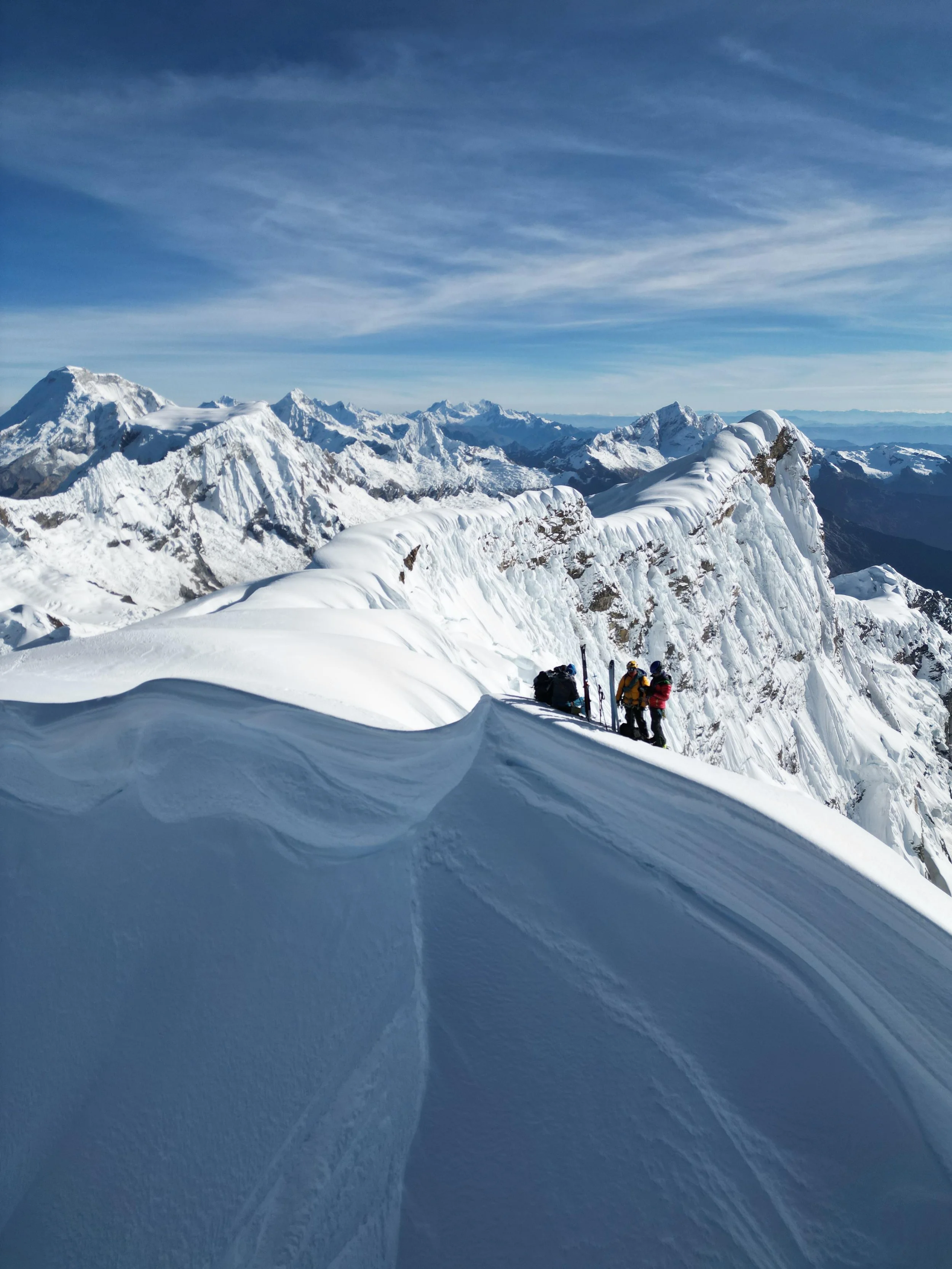 Snow-covered mountain ridge with a group of climbers resting on the edge, vast snow-capped peaks in the background, under a partly cloudy sky.