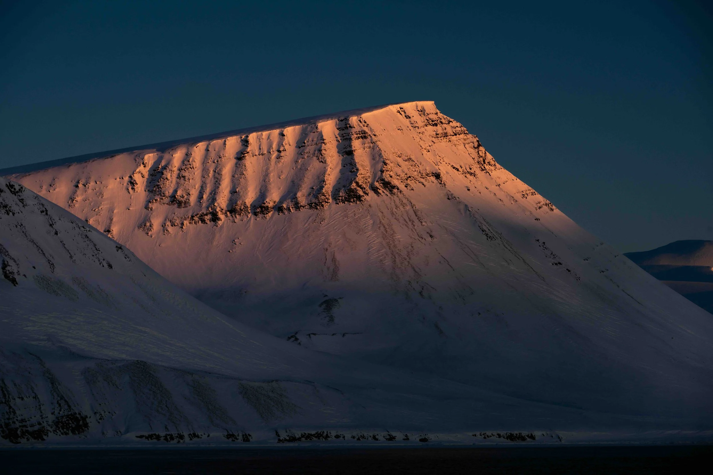 Snow-covered mountain at sunset with orange glow on the peak and dark sky in background.