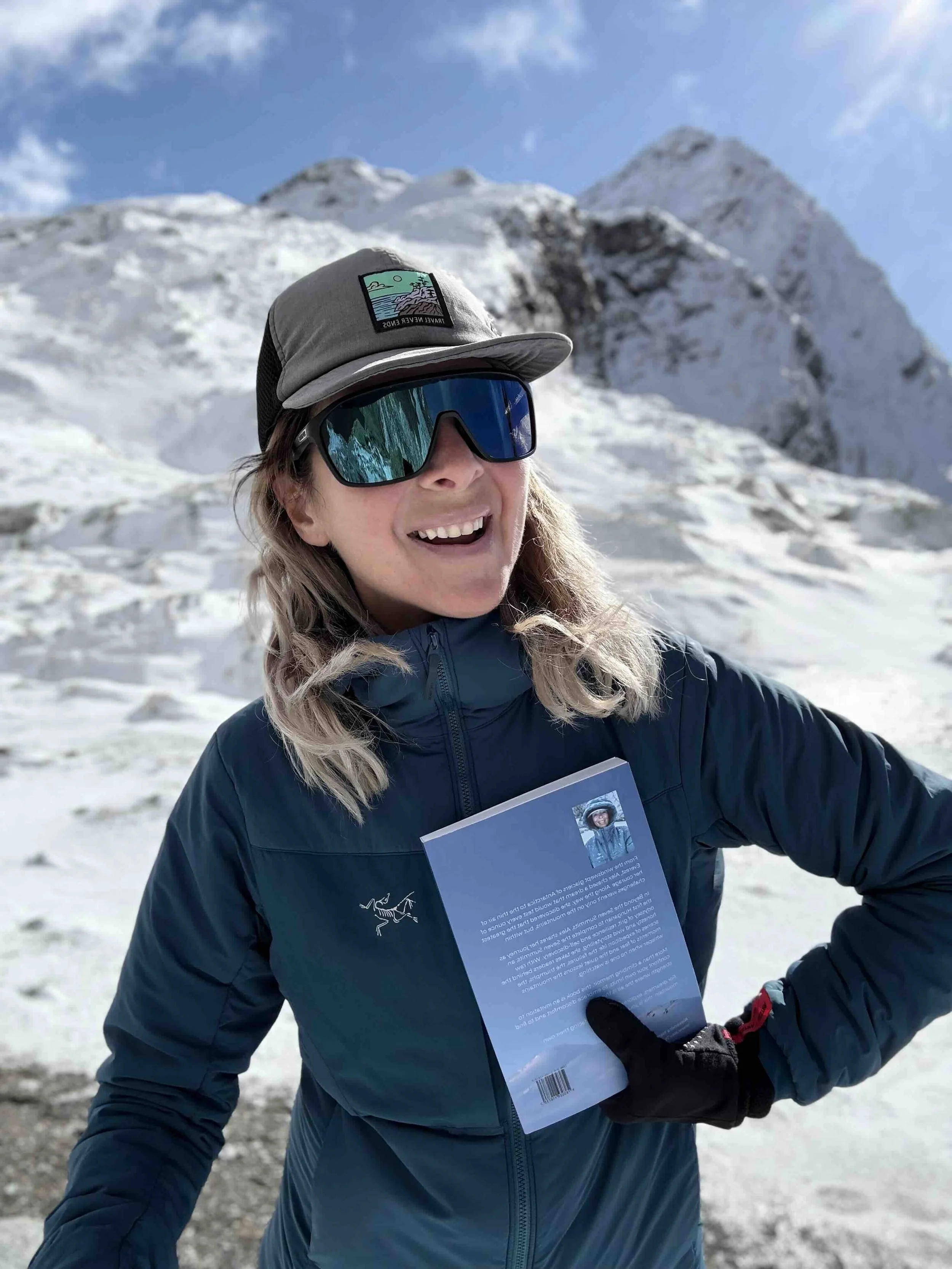 Me, Alex, standing in snowy mountains, smiling, wearing a blue Arc'teryx jacket, and black gloves, holding my recently published book, entitled Beyond the Seven Summits, Finding strength where the air is thin.