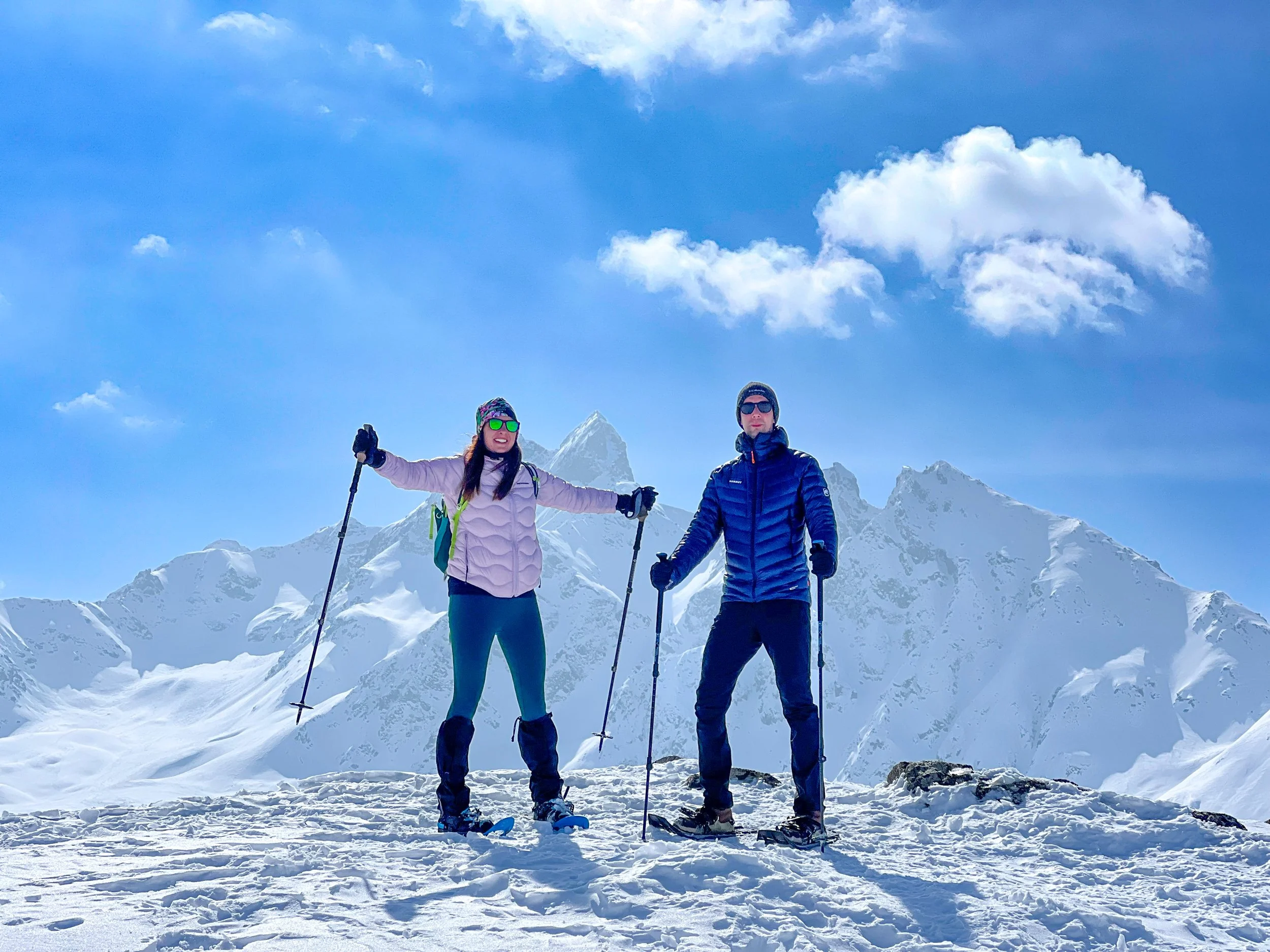 Two people standing on snow-covered mountain with snow-capped peaks in the background, wearing winter gear and holding ski poles, enjoying a snowshoe tour on Muottas Muragl Mountain in St. Moritz it the Swiss Alps, led privately by Alex.