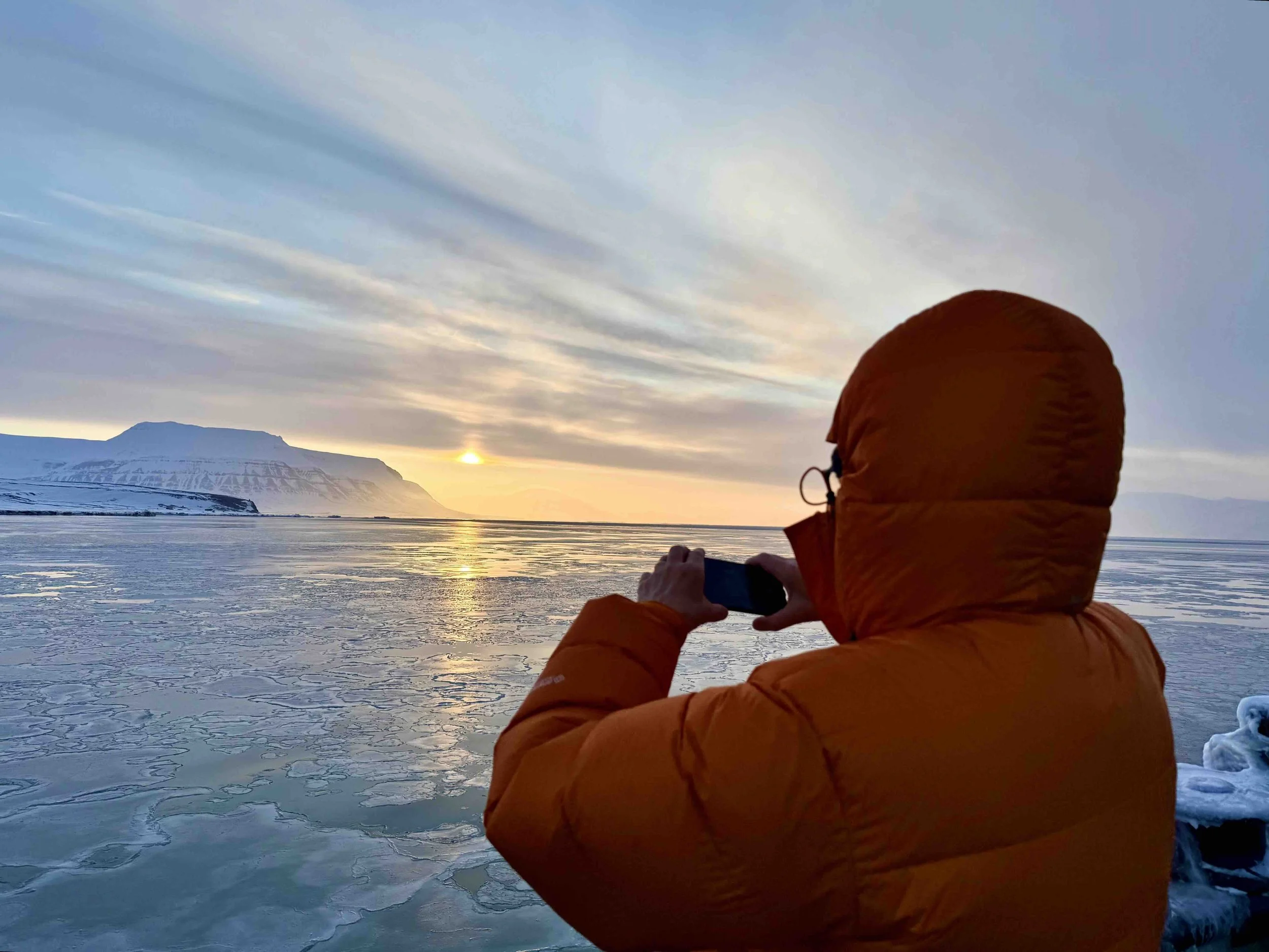 Person in an orange jacket taking a picture of a sunset over icy water with a mountain in the background.