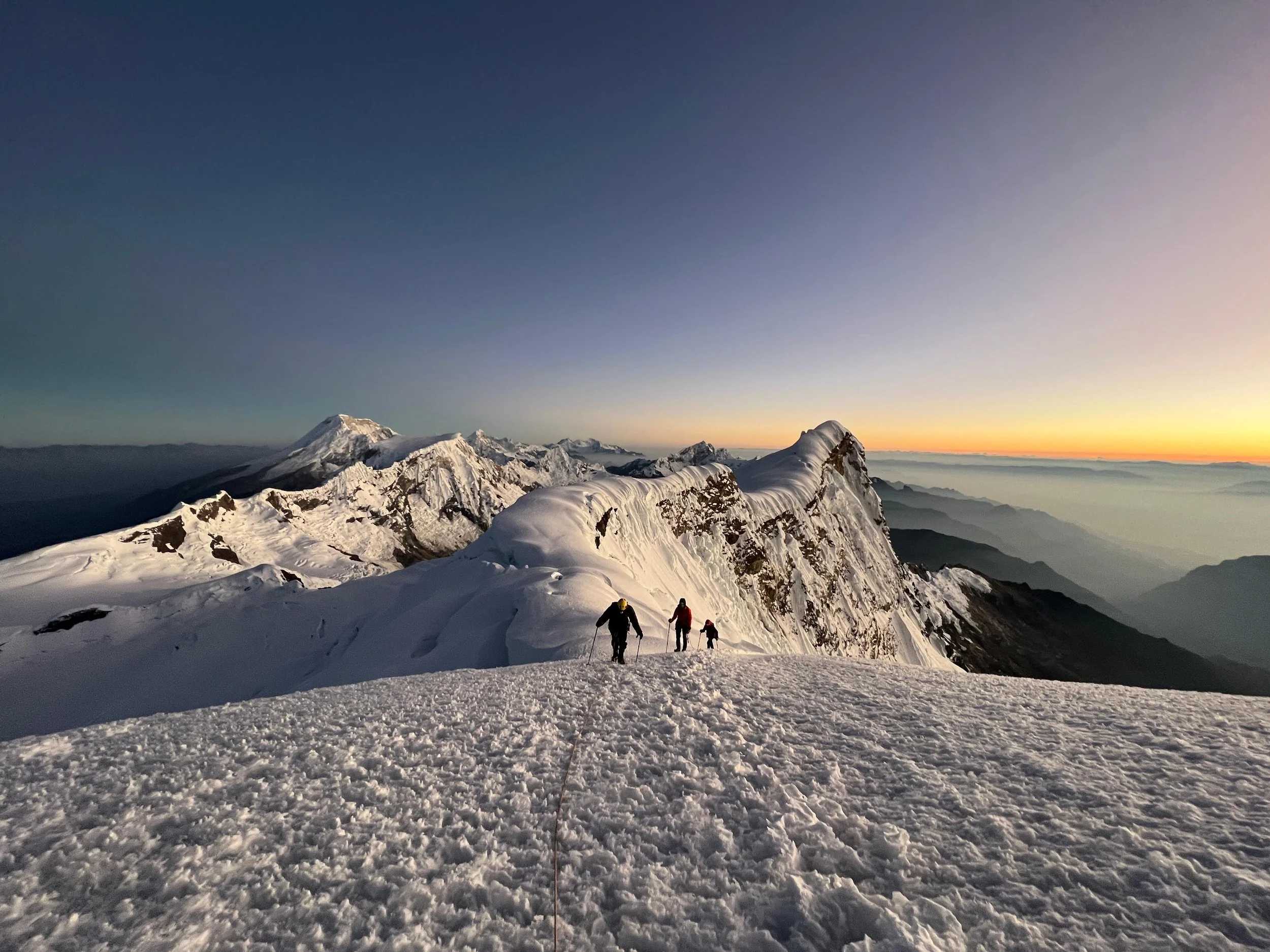 Snow-covered mountain peaks and climbers ascending a snowy ridge during sunrise in the Cordillera Blanca in Peru. Mountaineering expedition led by Alex.