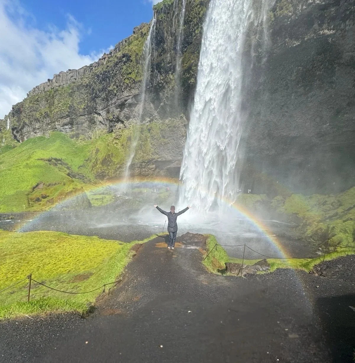 Brunette NYC escort and travel companion, Piper Leigh, raises her arms above her head underneath a waterfall and rainbow in Iceland
