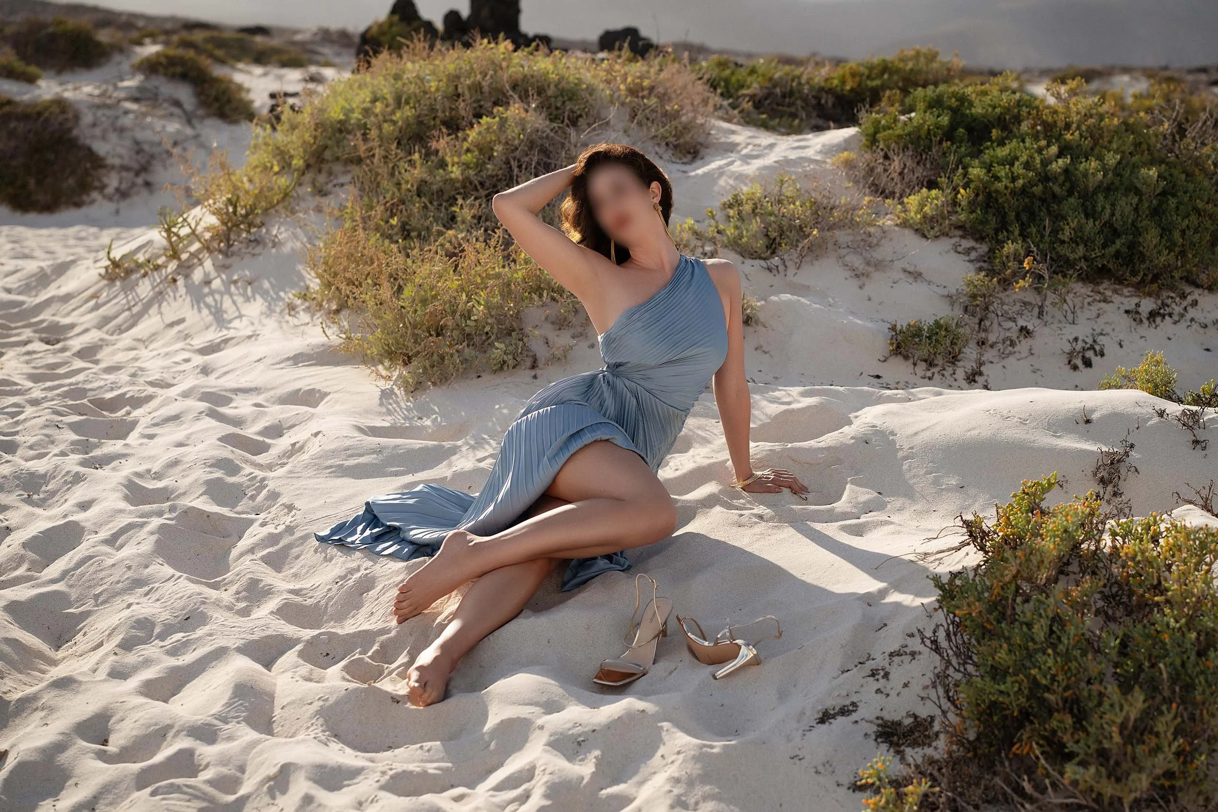 Brunette NYC escort, Piper Leigh, poses on a white sand beach in the Canary Islands wearing a long, pale blue dress