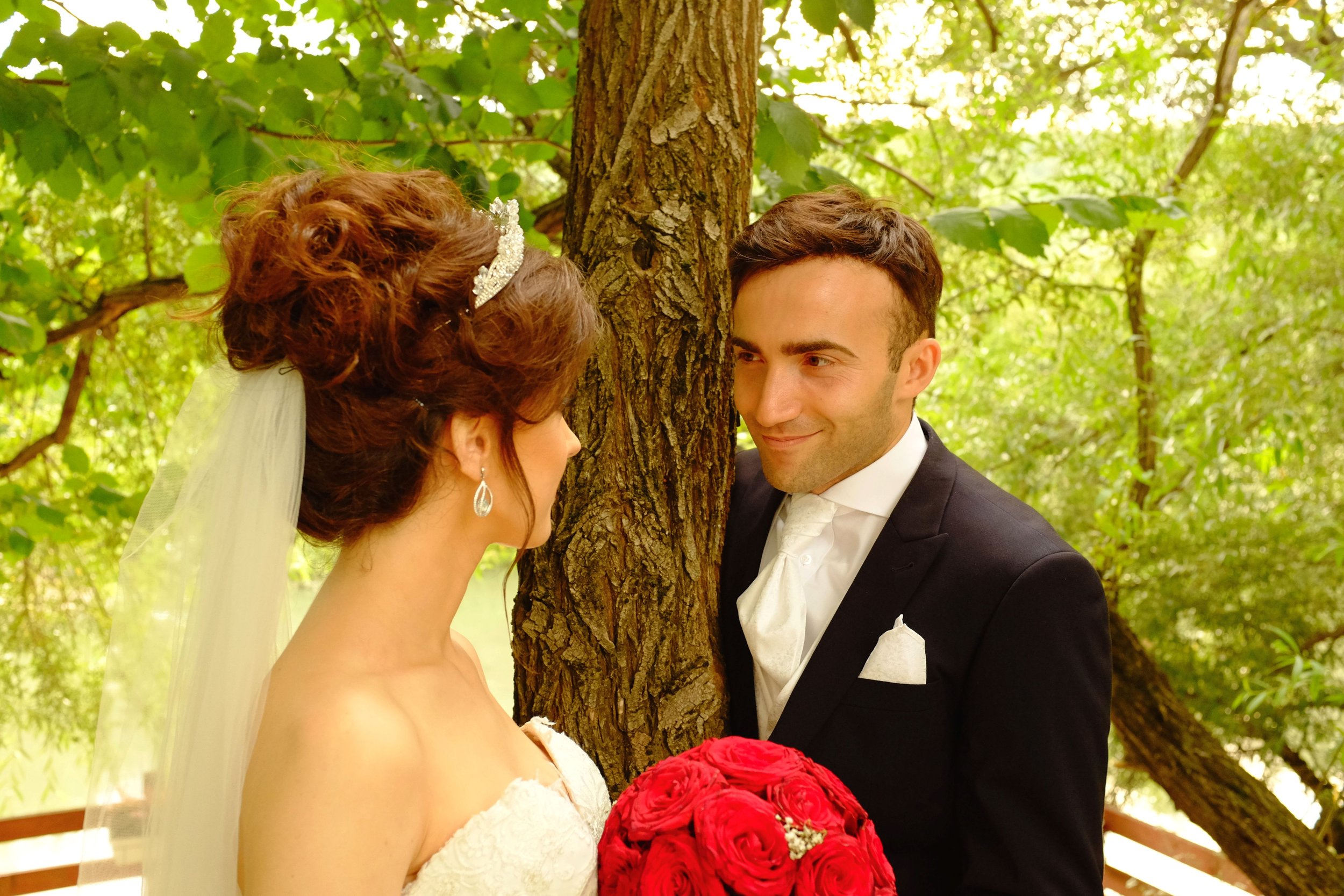 Ein Brautpaar im Hochzeitsoutfit steht sich gegenüber in einem grünen Wald. Die Braut trägt ein weißes Kleid, eine Tiara und eine Schleier, während der Bräutigam einen schwarzen Anzug mit einer weißen Krawatte und Einstecktuch trägt. Er hält einen roten Rosenstrauß. Sie schauen sich verliebt an.