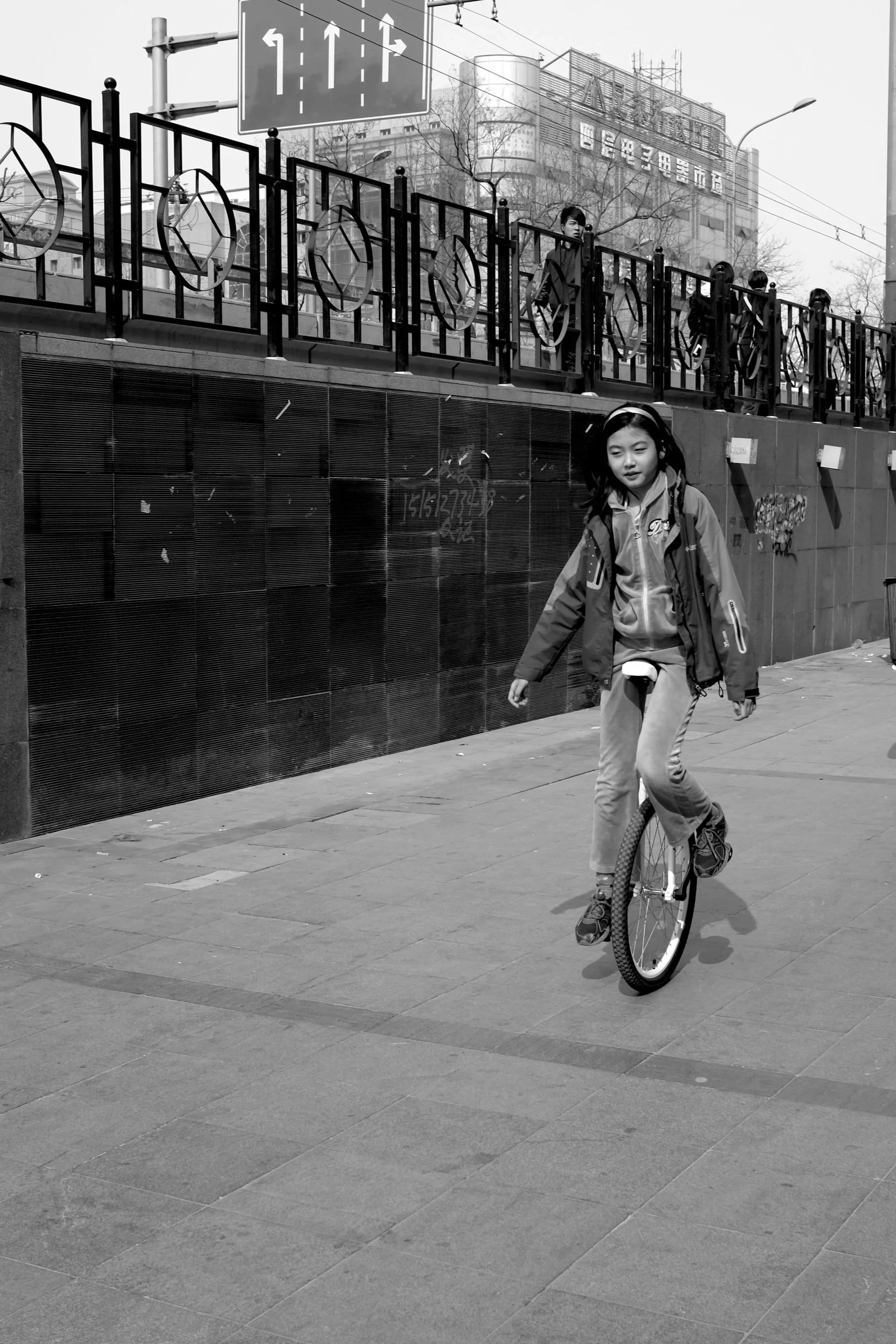 Eine Schwarz-Weiß-Aufnahme aus einer niedrigen Perspektive. Ein junges Mädchen fährt konzentriert auf einem Einrad über einen gepflasterten Gehweg. Im Hintergrund sind unscharf Passanten und urbane Strukturen einer chinesischen Großstadt zu erkennen.