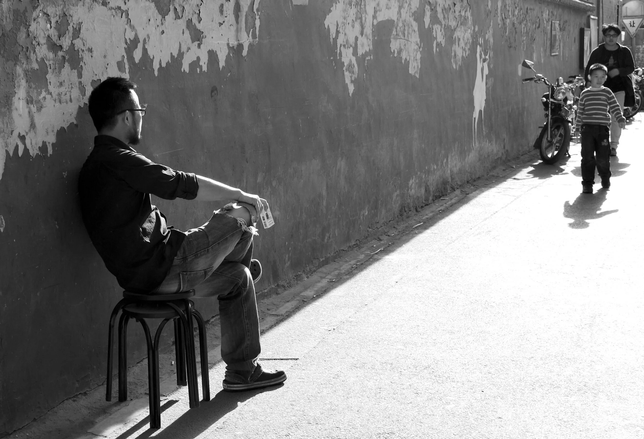 Schwarz-Weiß-Fotografie einer Straßenszene in einer Pekinger Gasse. Ein Mann sitzt entspannt auf einem kleinen Hocker und lehnt an einer hellen, texturierten Wand. Hartes Sonnenlicht fällt von der Seite ein und erzeugt tiefe, grafische Schatten.