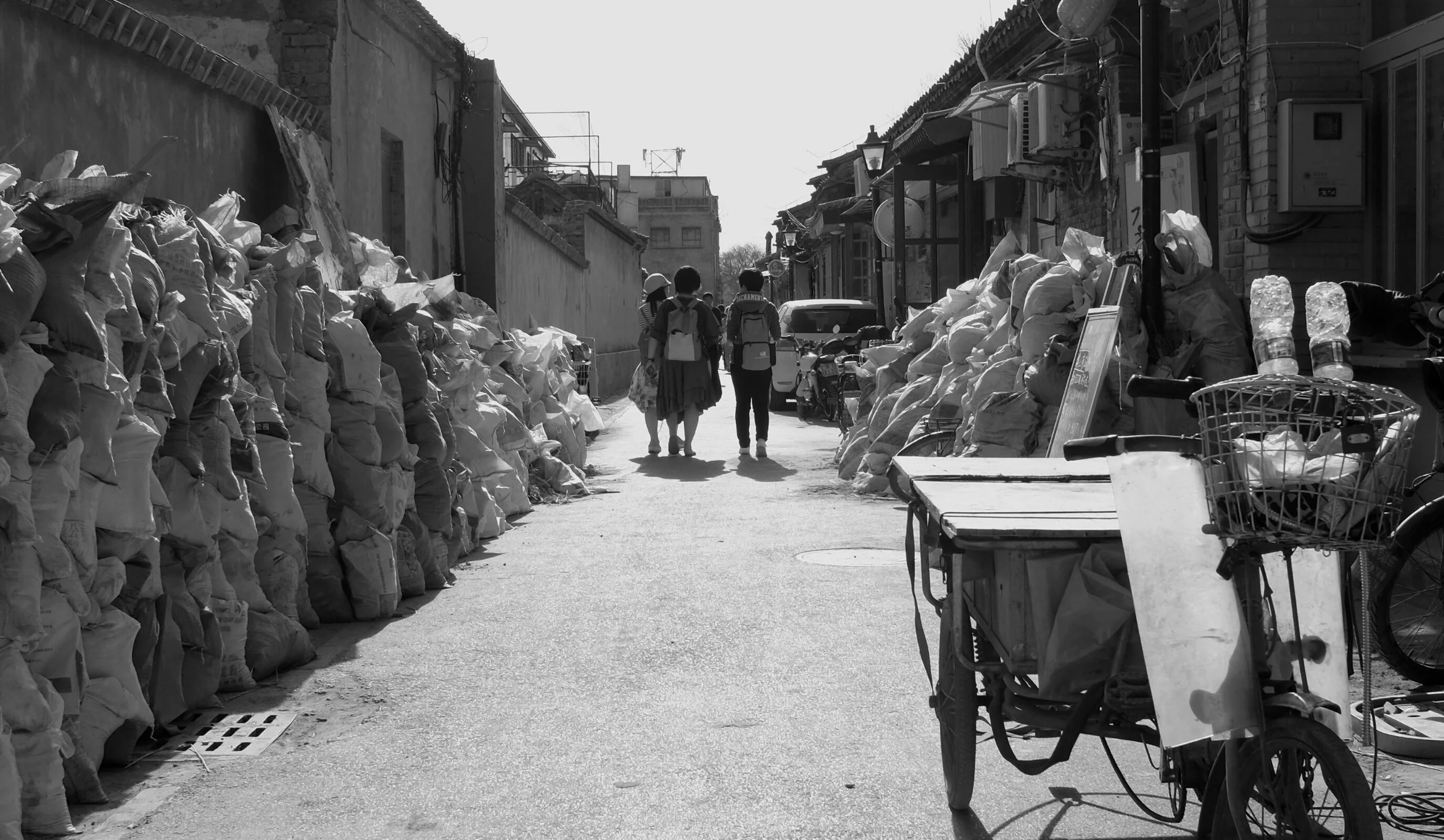 Fluchtpunkt-Perspektive einer schmalen Pekinger Gasse, flankiert von aufgetürmten Sandsäcken. Passanten im Hintergrund verleihen der kargen, grafischen Szene Leben. Ein minimalistisches Werk aus der Selektion von pvre u, das Texturen und Linien beton