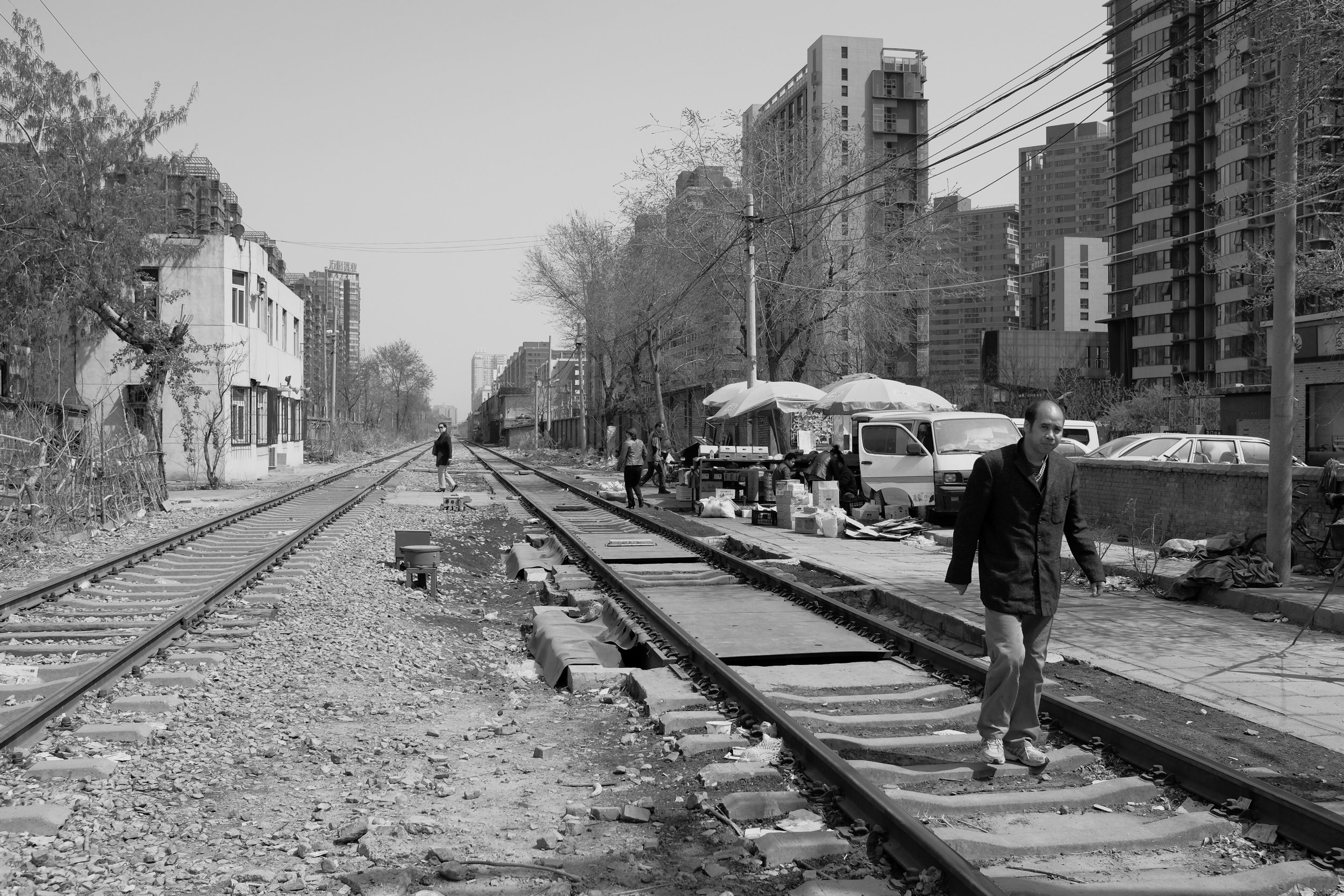 Urbane Kontraste in Peking: Marktstände direkt an einer aktiven Gleisanlage, gesäumt von modernen Wohnblöcken. Die weite Perspektive und das klare Tageslicht unterstreichen den dokumentarischen Charakter dieser pvre u Aufnahme.