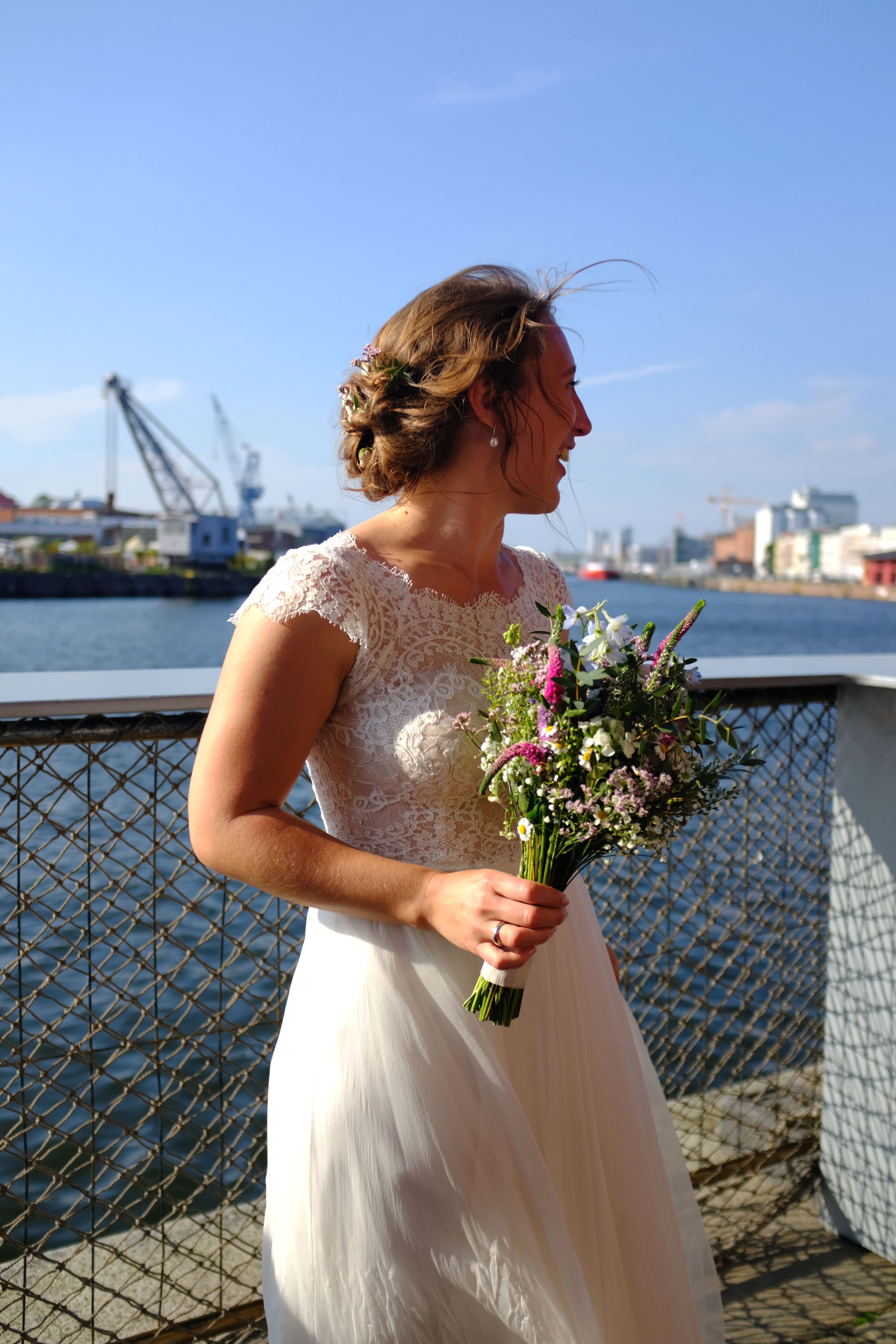 Eine Braut in einem weißen Kleid hält einen Blumenstrauß und steht an einem Wasserweg mit Hafen und Gebäuden im Hintergrund, bei sonnigem Himmel.