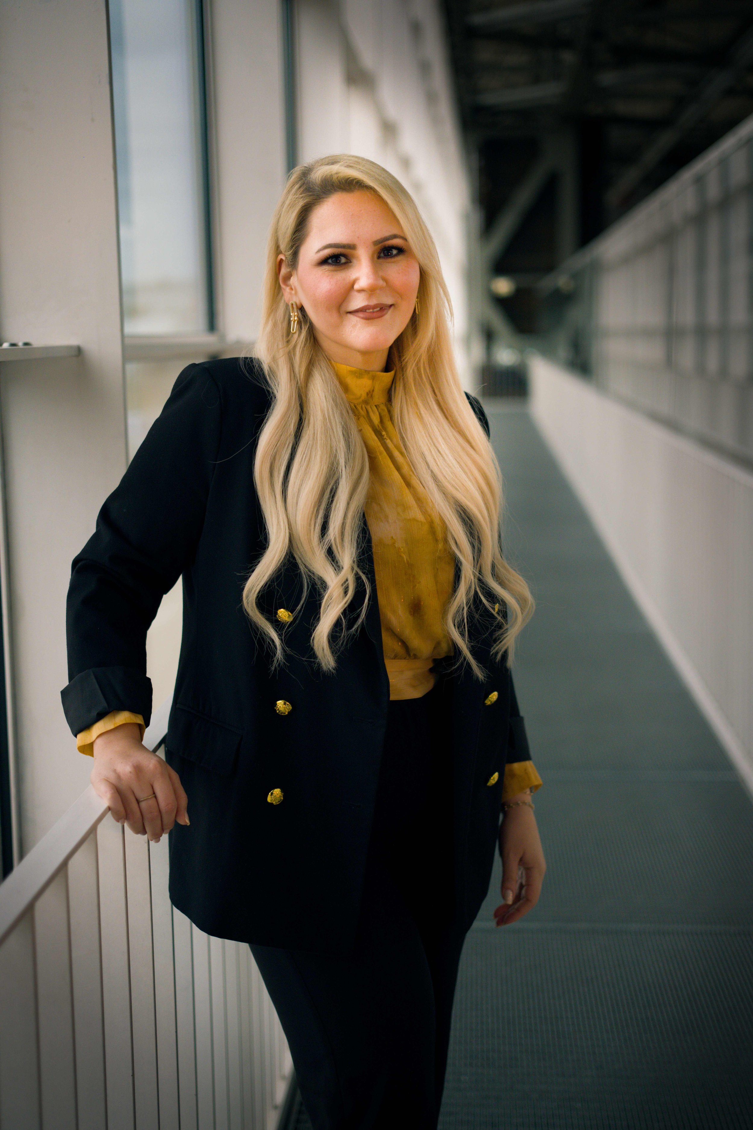 Eine junge Frau mit langen blondem Haar, schwarze Jacke mit goldenen Knöpfen, gelbem Seidenbluse, steht an einem Gang in einem modernen Gebäude, lächelnd in die Kamera schauend.