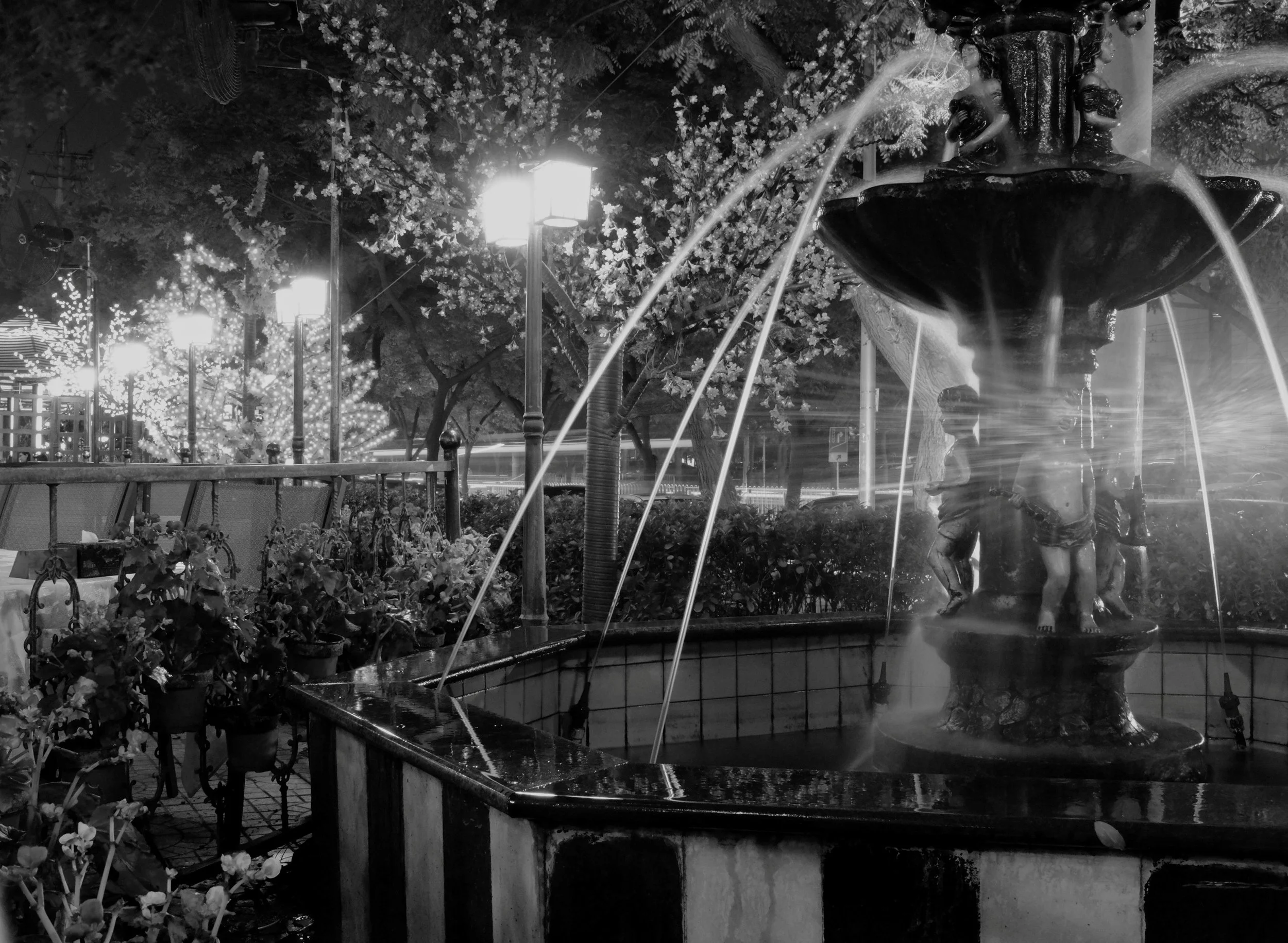 Nächtliche Straßenszene in Peking mit einem kunstvollen Springbrunnen im Fokus. Weiches Licht historischer Straßenlaternen kontrastiert mit den harten Schatten der Umgebung. Authentische Schwarz-Weiß-Fotografie von pvre u, die Ruhe und urbane Ästheti