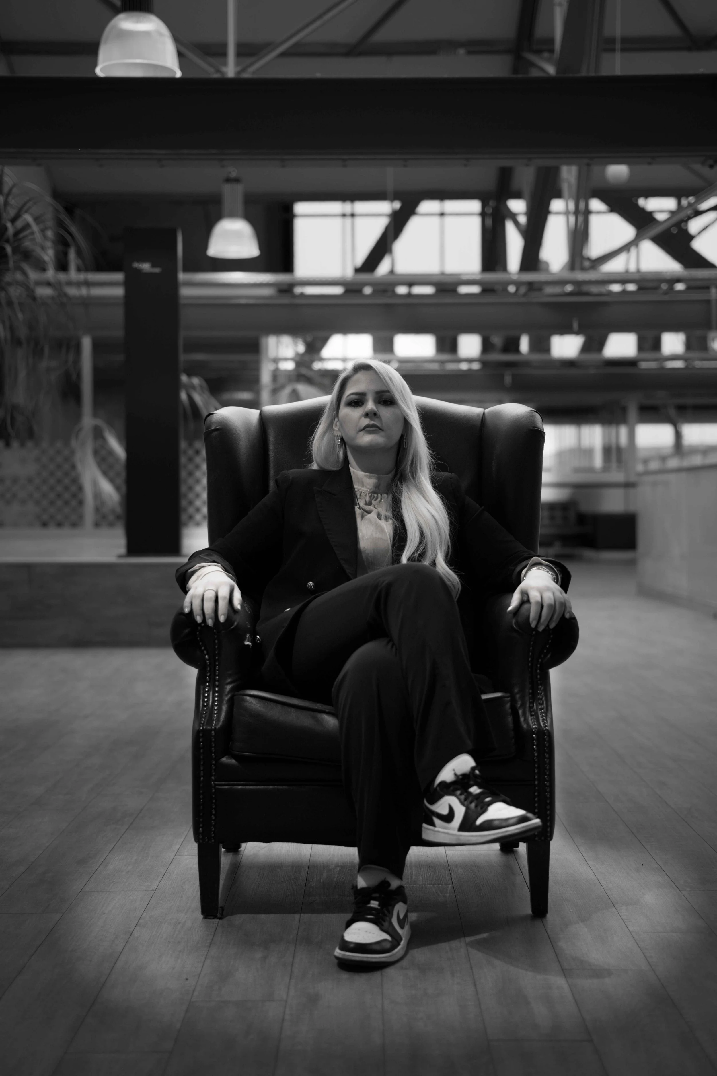 Schwarz-Weiß-Bild einer selbstbewussten Frau, die in einem großen Ledersessel sitzt, in einem modernen, industrial-stil Innenraum, trägt einen schwarzen Anzug und Nike-Sneaker, wirkt ernst und entspannt.