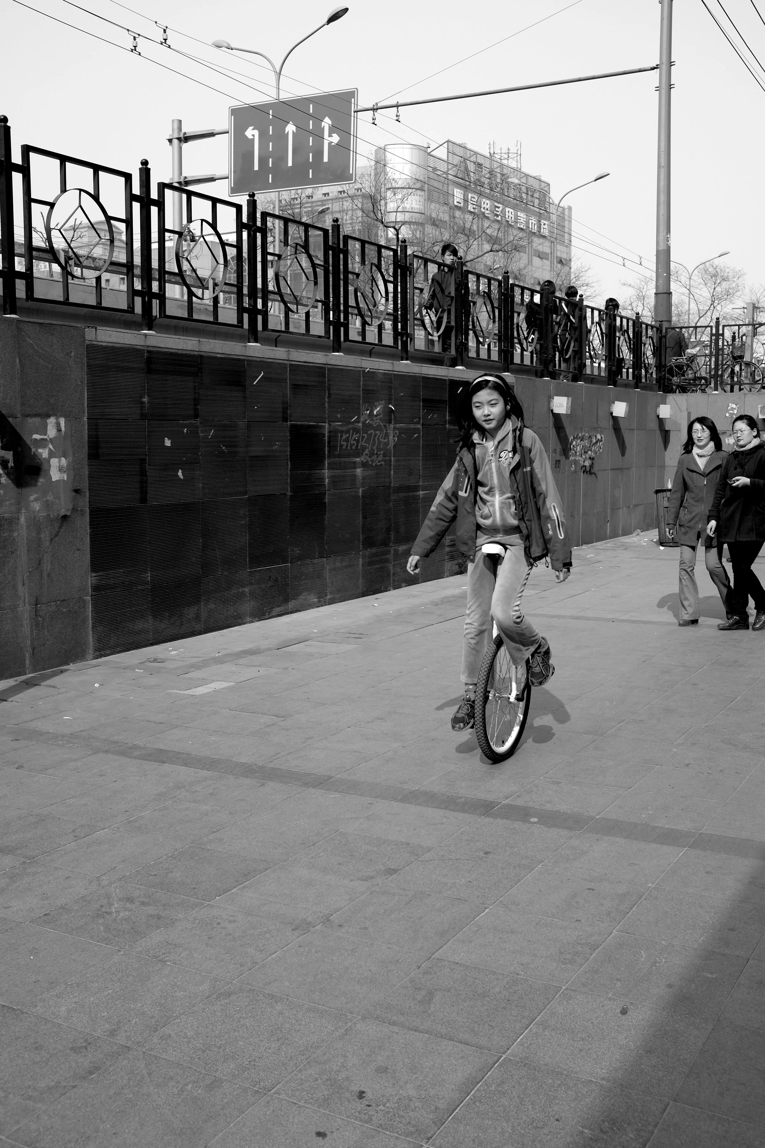 Dynamischer Moment auf einem Pekinger Gehweg: Ein junges Mädchen balanciert geschickt auf einem Einrad. Die Schwarz-Weiß-Fotografie fängt die Bewegung und Konzentration vor einer urbanen Kulisse im typischen pvre u Stil ein.