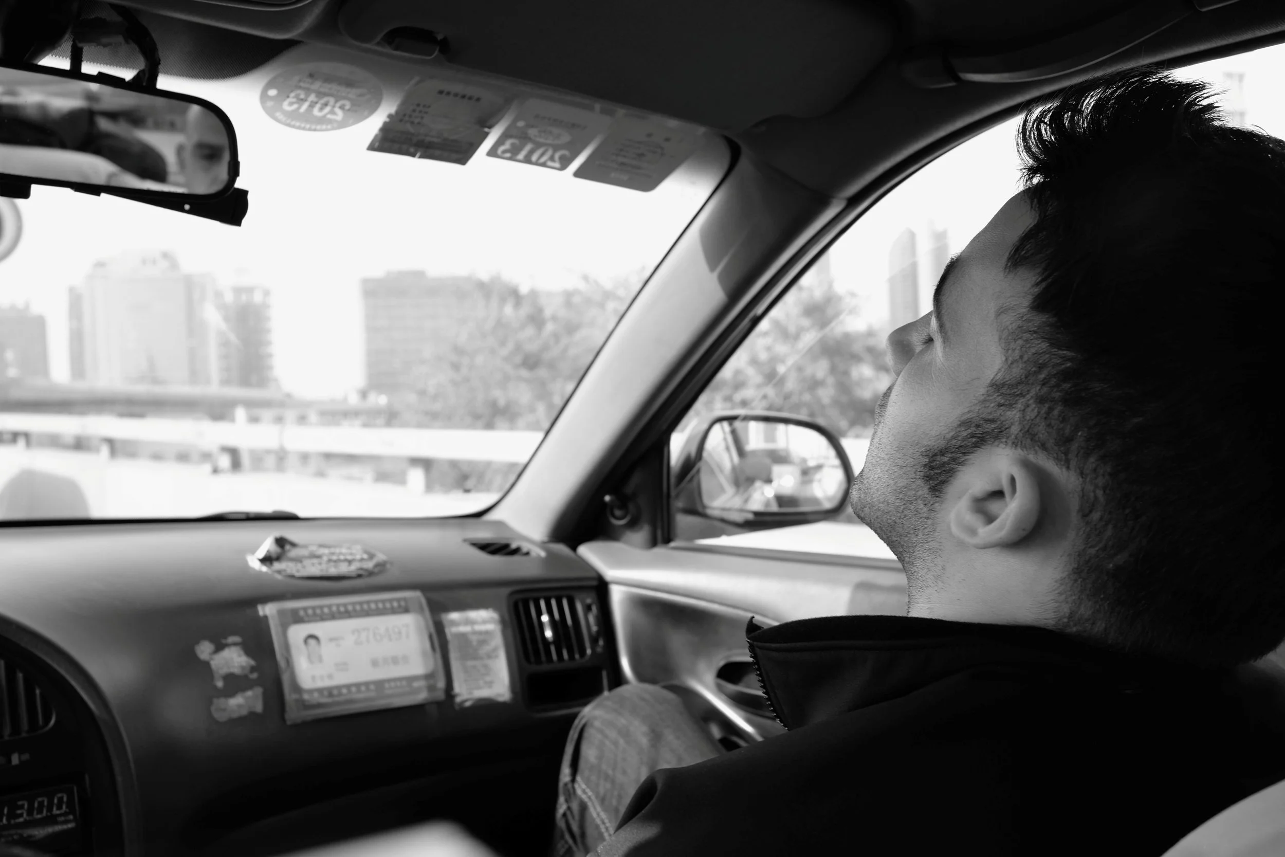 Ein junger Mann sitzt im Auto, der Blick nach rechts gerichtet, im Vordergrund die Fahrzeugfront und der Rückspiegel. Im Hintergrund sind Stadtgebäude sichtbar.