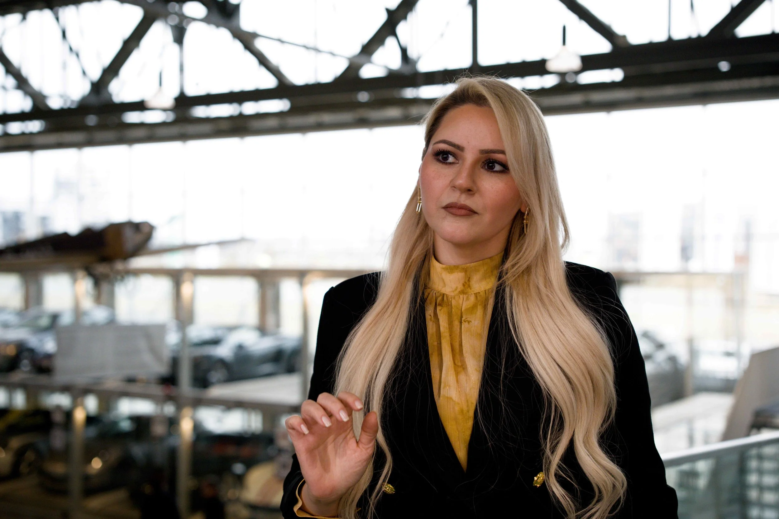 Eine blonde Frau trägt einen schwarzen Blazer und ein gelbes Top, steht in einem Parkhaus, im Hintergrund sind geparkte Autos sichtbar.