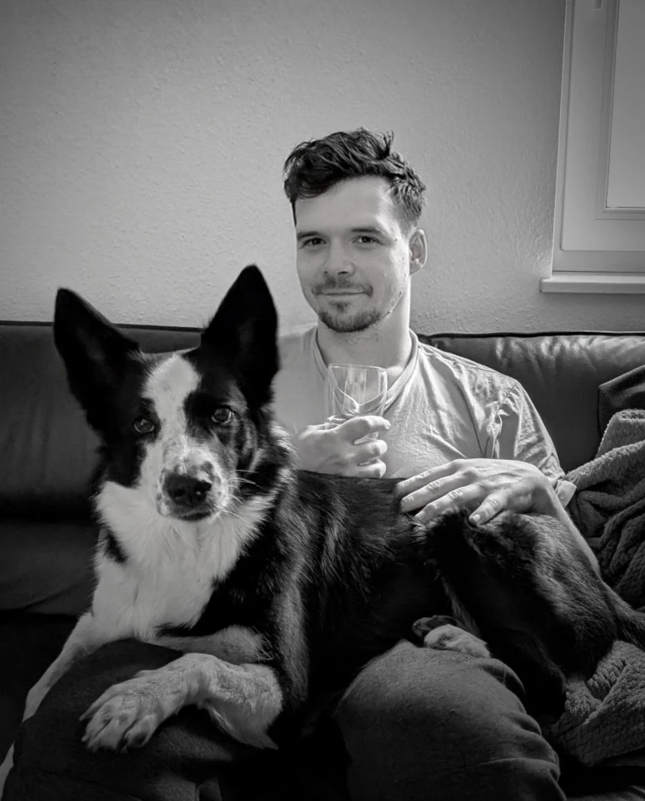 Ein Mann sitzt auf einer Couch und hält ein Weinglas, neben ihm liegt ein Hund mit schwarz-weißer Fellzeichnung, beide betrachten die Kamera.