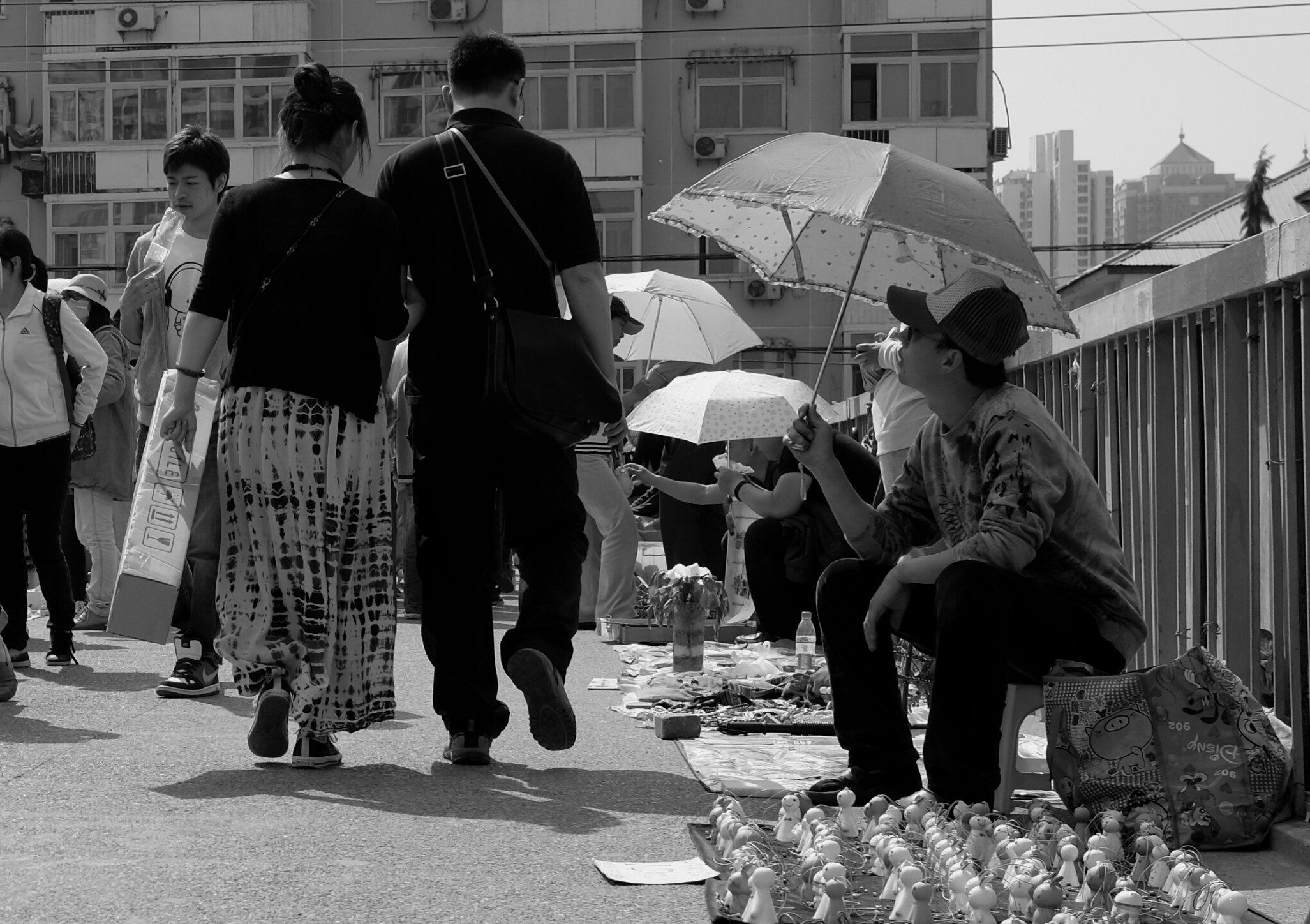 Menschenmenge und Straßenhändler auf einer Brücke in Peking unter Sonnenschirmen. Die Komposition spielt mit verschiedenen Ebenen und menschlicher Interaktion. Street-Photography von pvre u, die das soziale Gefüge der Stadt dokumentiert.
