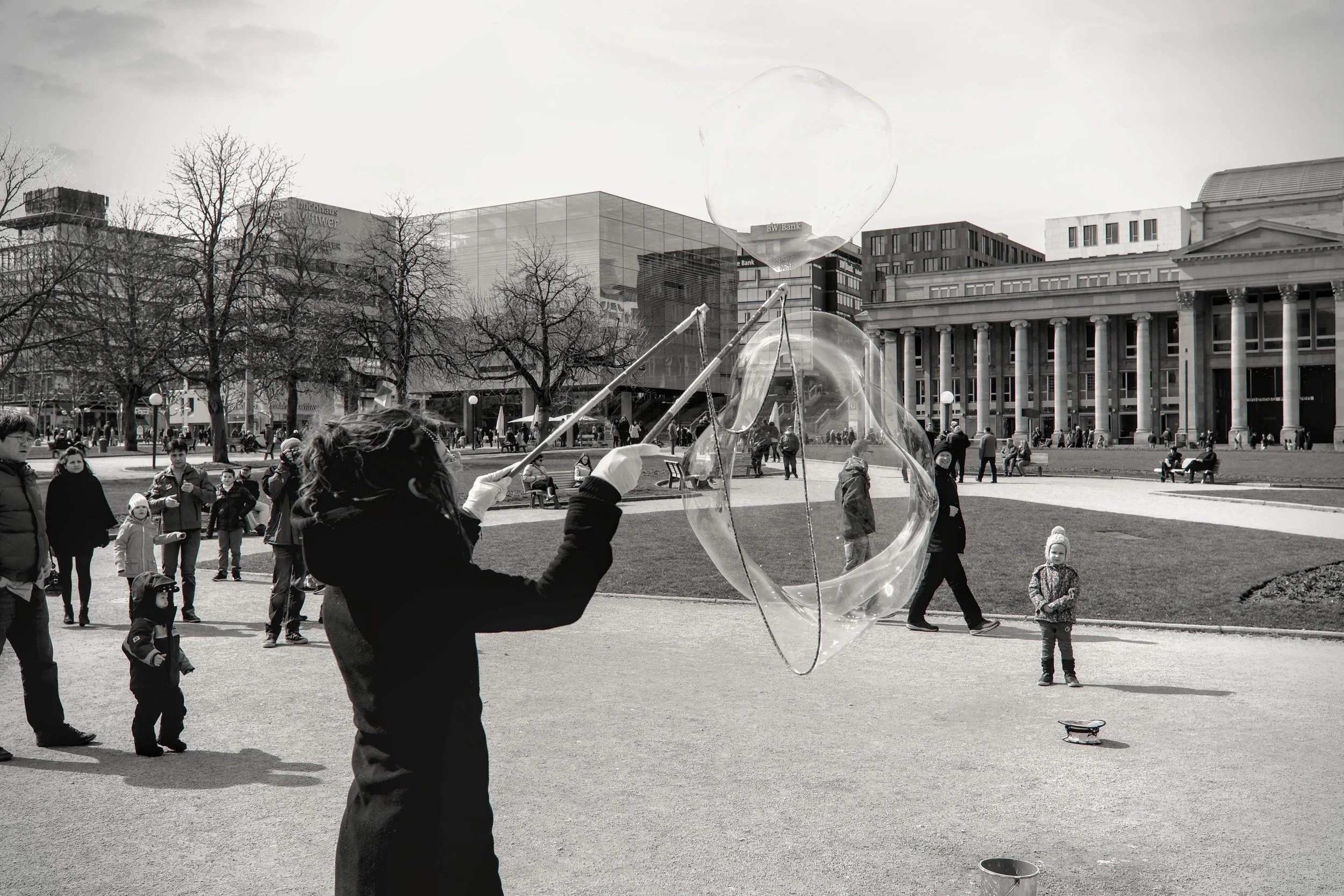 Eine Person, wahrscheinlich eine Straßenkünstlerin, macht eine große Seifenblase in einem Stadtpark. Kinder und Erwachsene beobachten die Seifenblase, während sie in der Luft schwebt. Im Hintergrund sind Bäume, moderne Gebäude und ein Theatergebäude sichtbar.