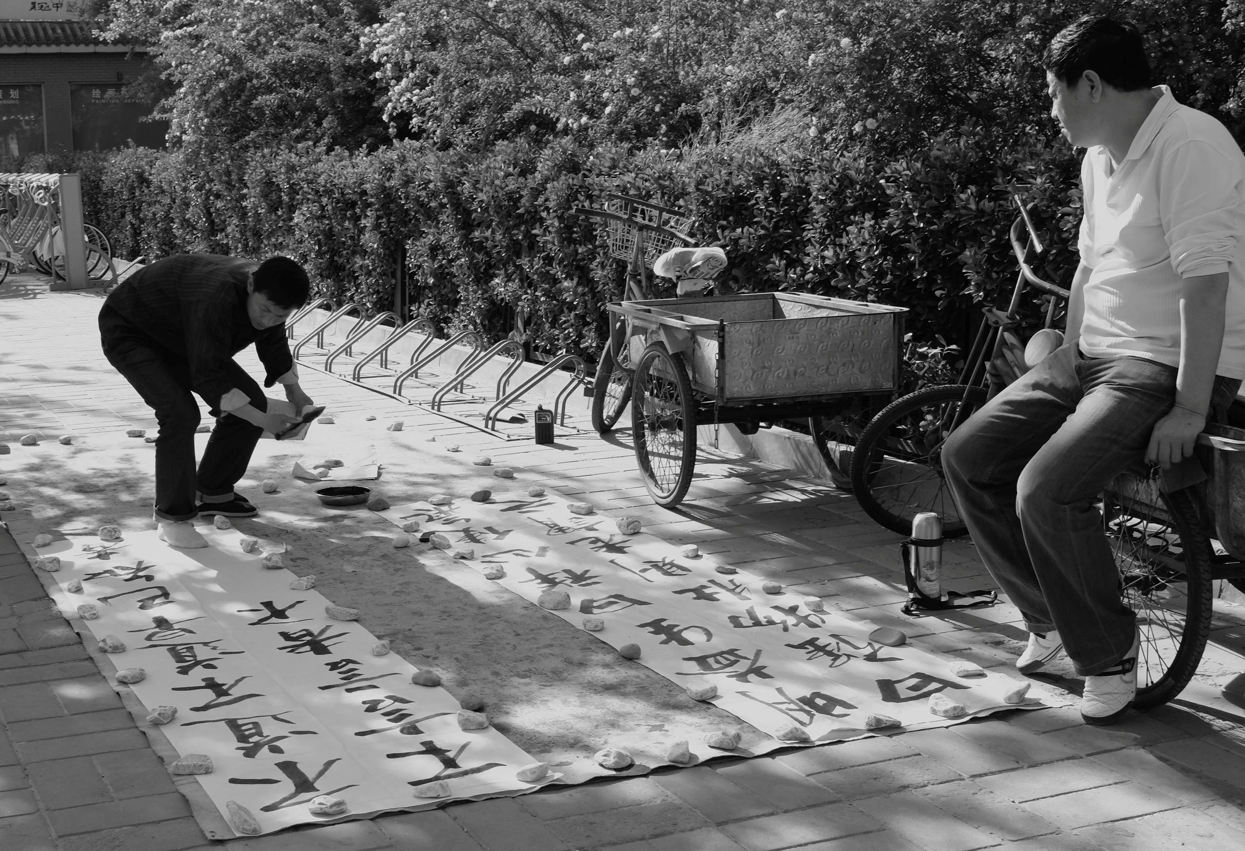 Schwarz-Weiß-Aufnahme eines Straßenkünstlers in Peking. Der Mann kniet am Boden und schreibt mit einem Pinsel Kalligraphie auf lange weiße Papierbahnen, die mit Steinen beschwert sind.  hartes Sonnenlicht betont die Kontraste auf dem Boden
