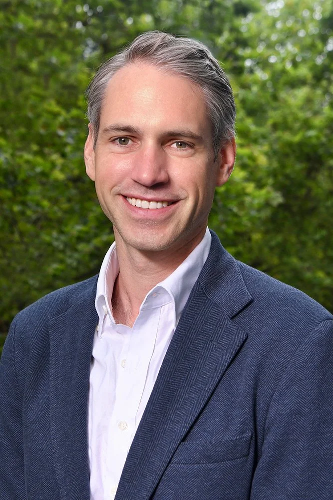 Portrait of a smiling man with gray hair, wearing a navy blazer and white shirt, standing outdoors with green foliage in the background.