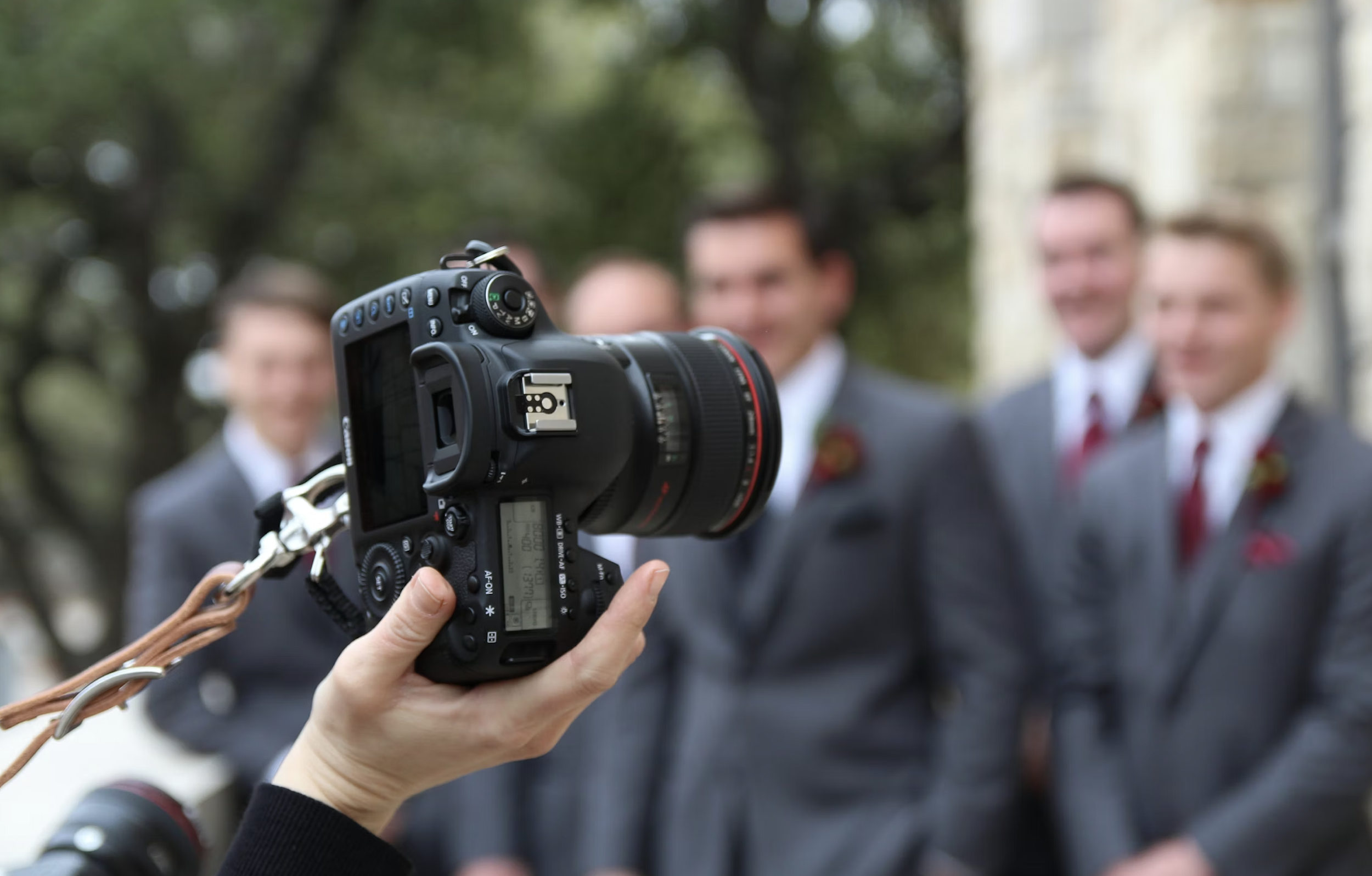 A person holding a professional digital camera in front of four men in suits, with the men being out of focus, outdoors with trees and a building in the background.