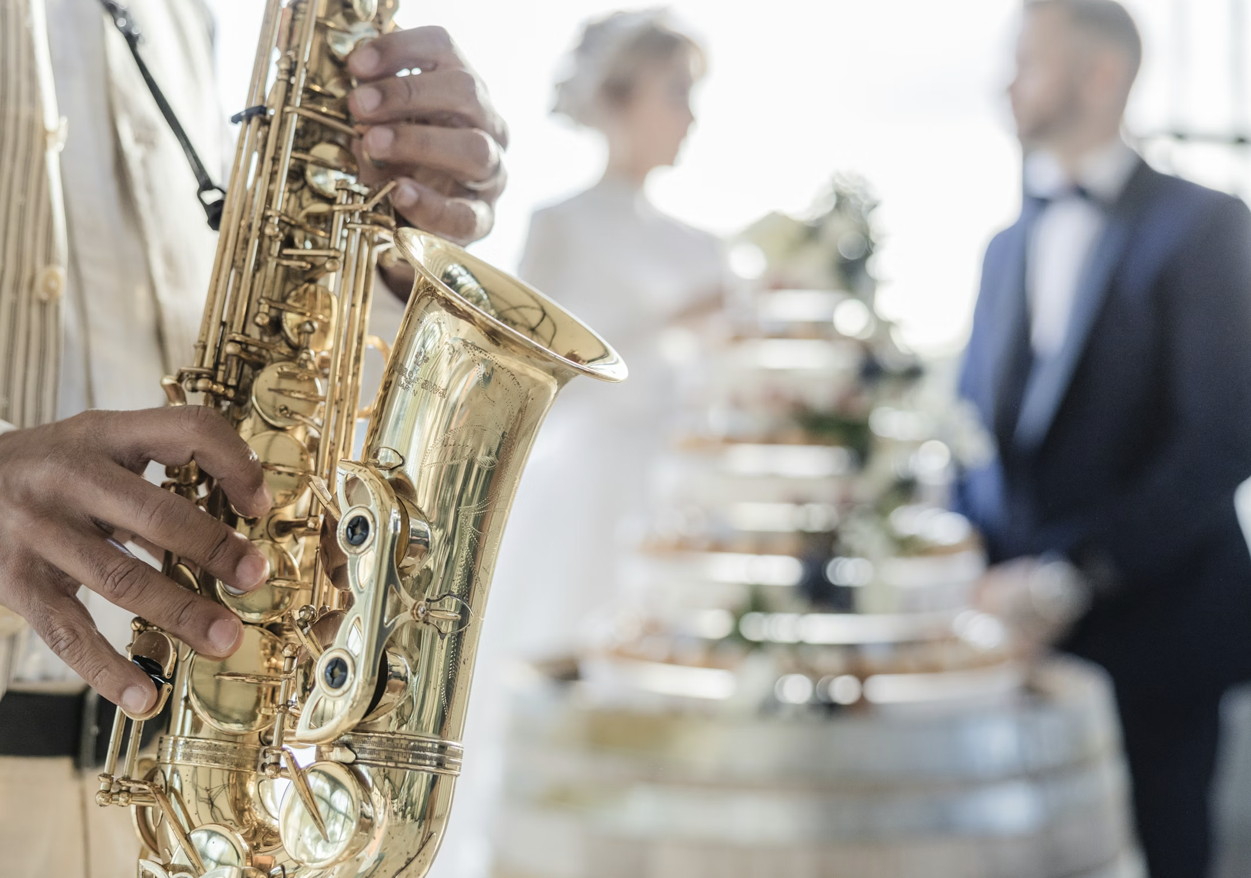 Close-up of a person's hands playing a gold saxophone during a formal event with a blurred couple in tuxedo and gown in the background.