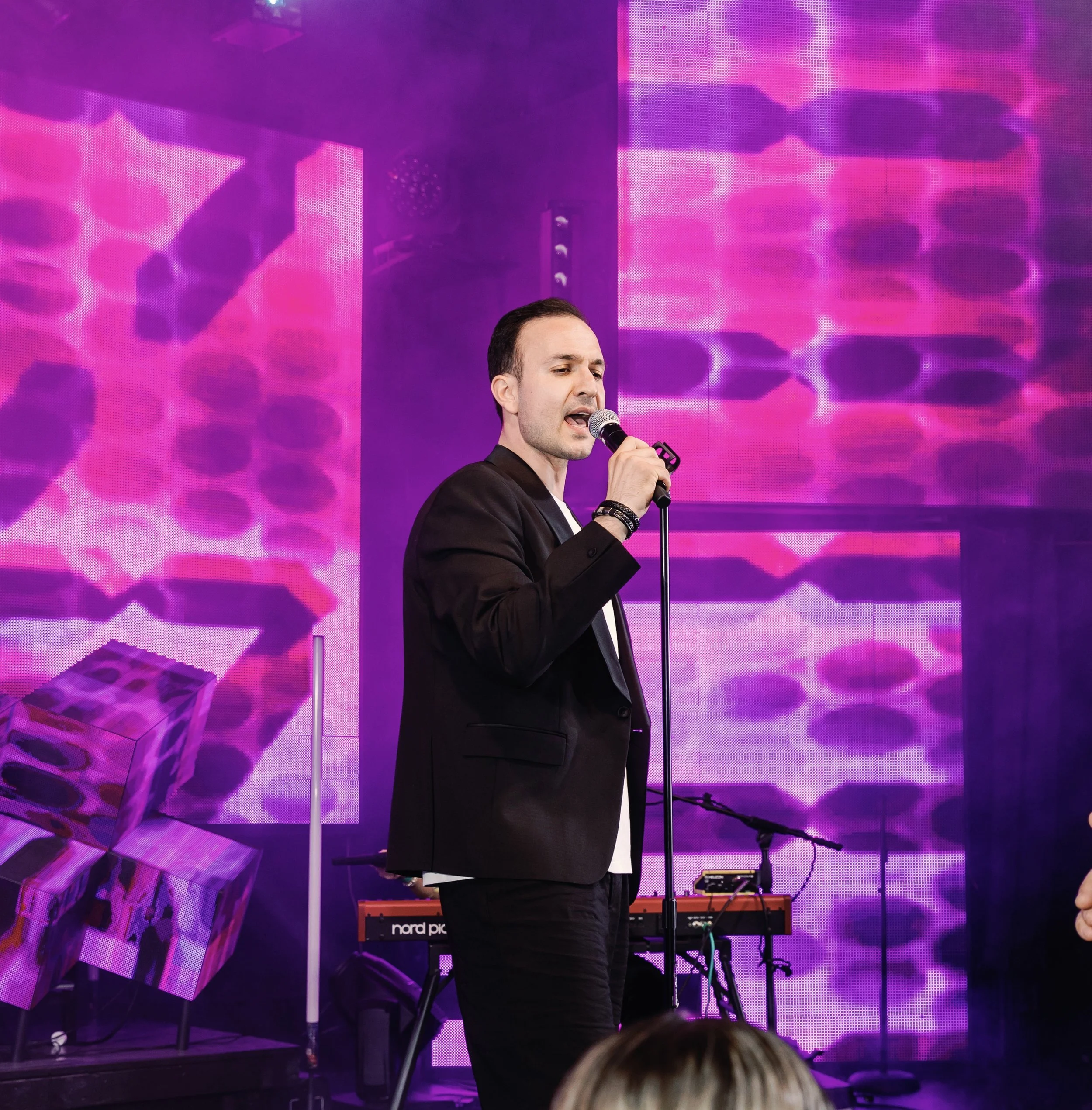 A man singing into a microphone on a stage with vibrant pink and purple LED screens in the background and musical equipment around him.