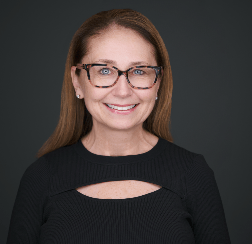 A smiling woman with reddish-brown hair, wearing glasses and small earrings, dressed in a black top with a keyhole cutout, against a dark gray background.