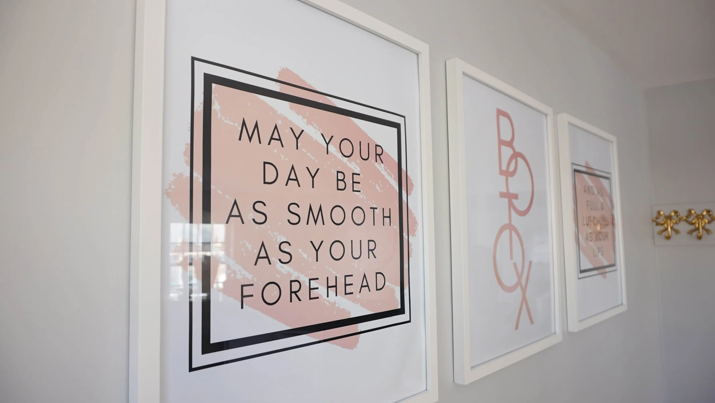 Decorative framed posters on a white wall, including one that says "May your day be as smooth as your forehead" with pink abstract background.