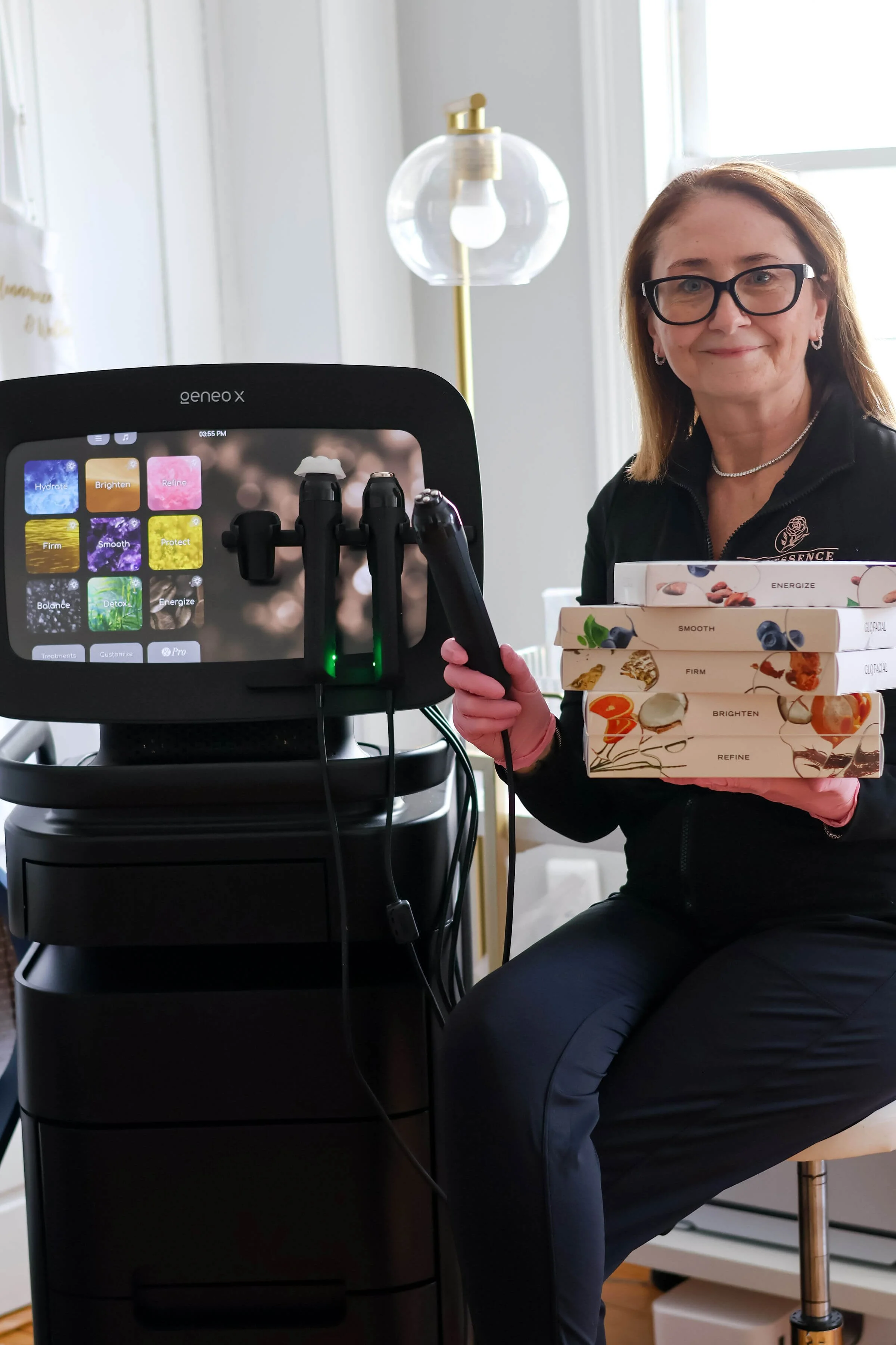A woman with glasses and a black jacket is sitting and smiling while holding multiple skincare product boxes in her hand. She is sitting next to a machine with a touchscreen display and various attachments.