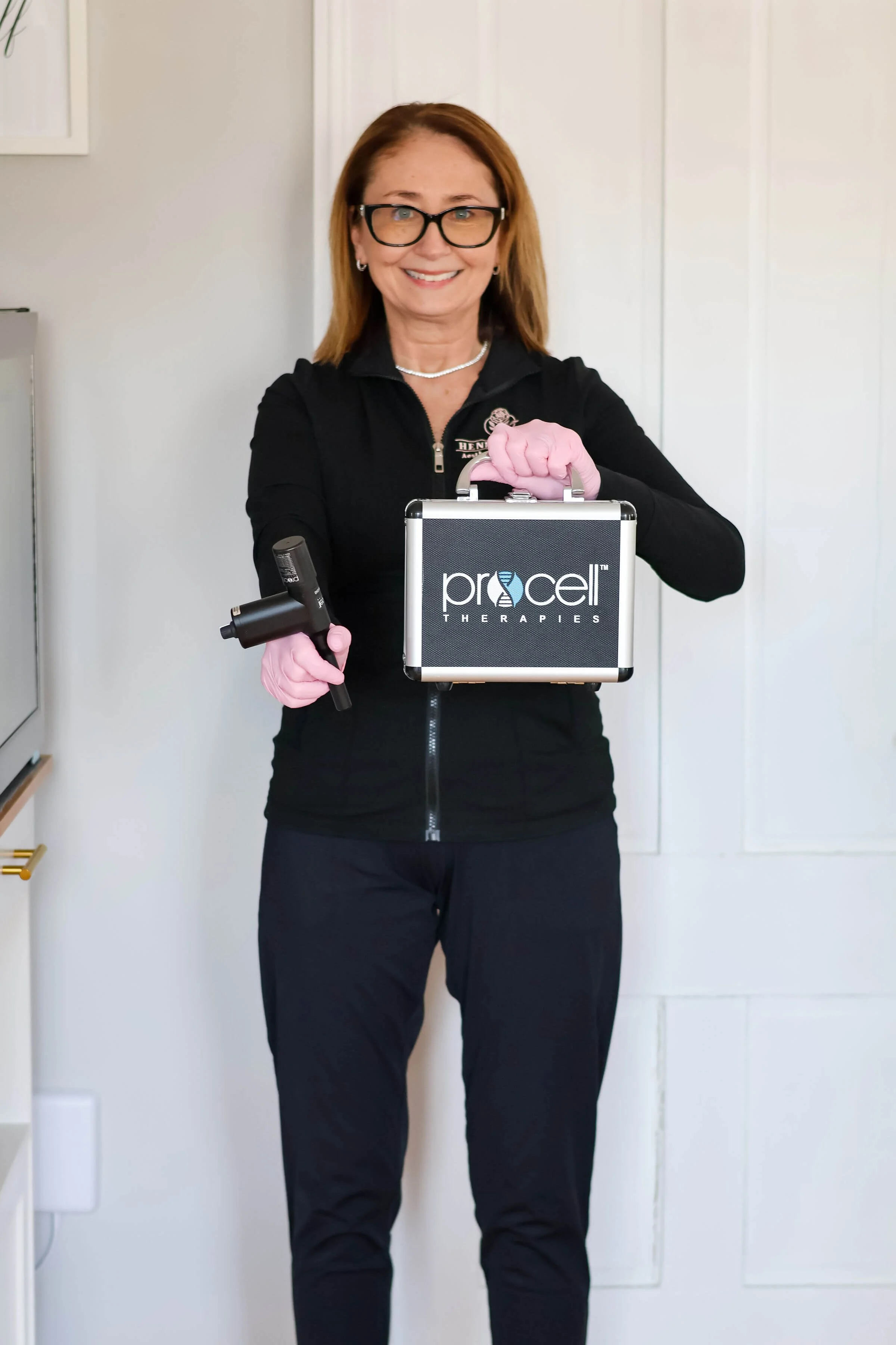 A woman wearing glasses, black clothing, and pink gloves is holding a small black case with the logo 'Procull Therapies' and a handheld device, standing indoors in front of a white wall.