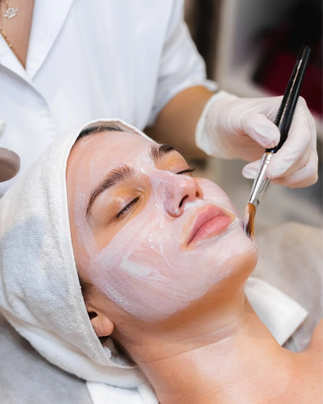 A woman receiving a facial treatment, with a beauty professional applying a facial mask using a brush.