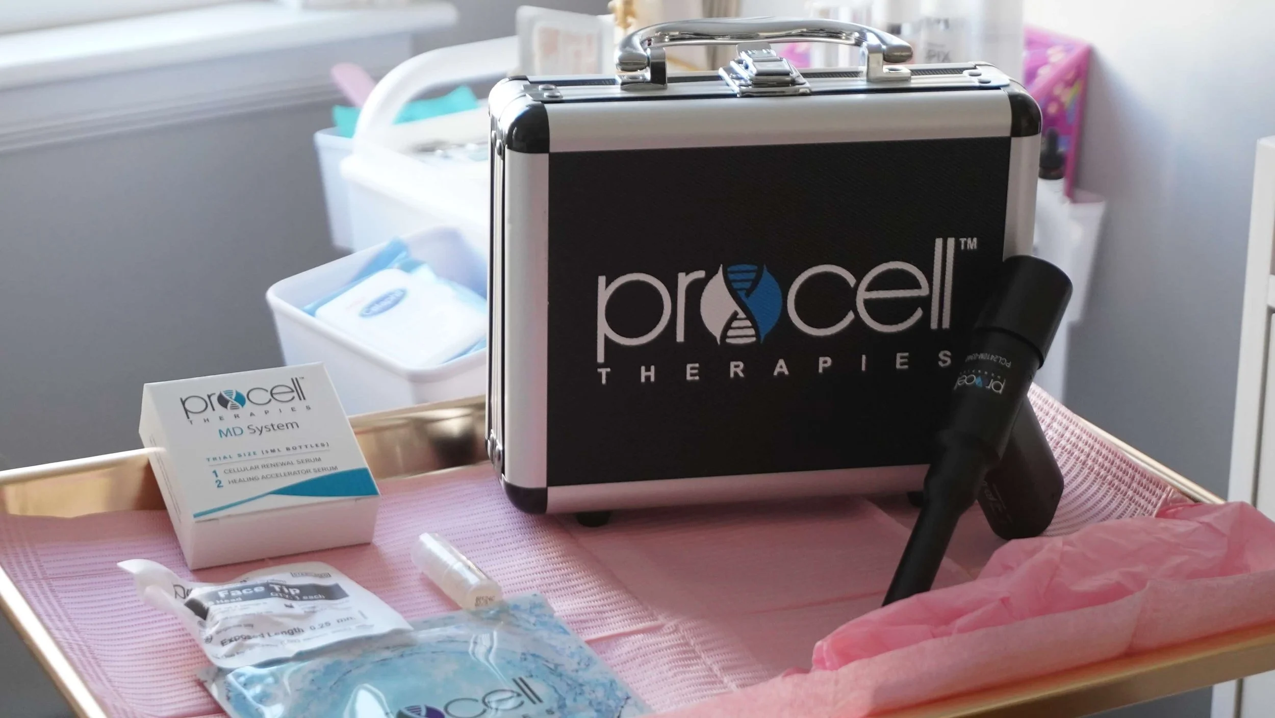 A medical treatment kit labeled 'Procella Therapies' on a pink tray, featuring a black and silver case, a box labeled 'Procella MD System', syringes, and other medical supplies.