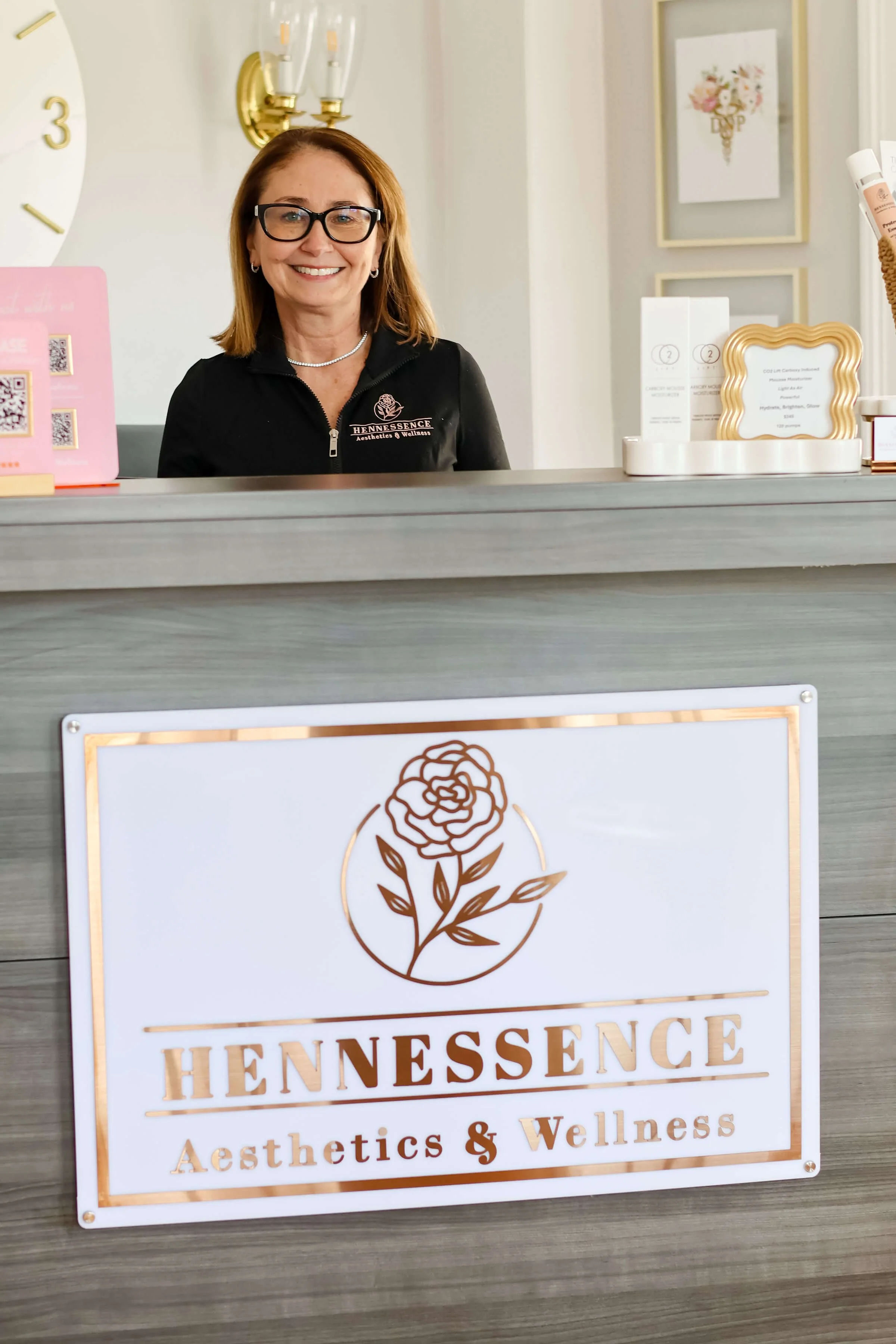 A smiling woman with glasses and shoulder-length hair standing behind a reception counter at Hennessence Aesthetics & Wellness. The reception desk has a sign with a rose logo and the business name, with several framed art pieces in the background.