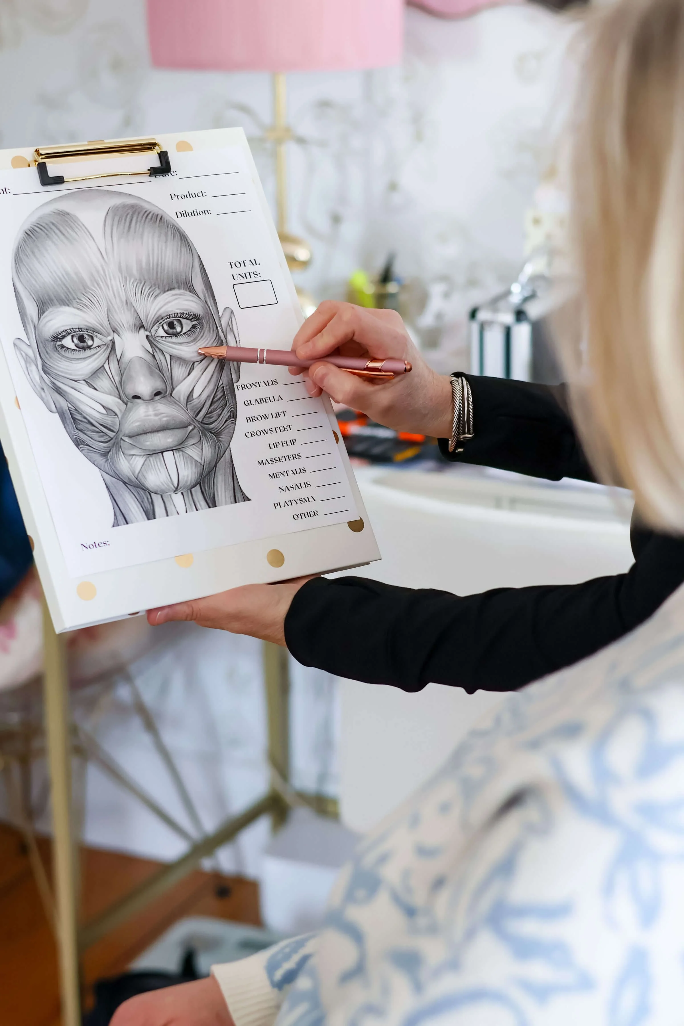holding a chart of the human face with detailed facial muscles, showing areas labeled such as frontalis, glabella, brow lift, crows feet, lip flip, masseters, mentalis, nasalis, platysma, and other. The artist is using a pen to point at the chart. The setting appears to be a tattoo studio with a backdrop of a wall and some equipment.