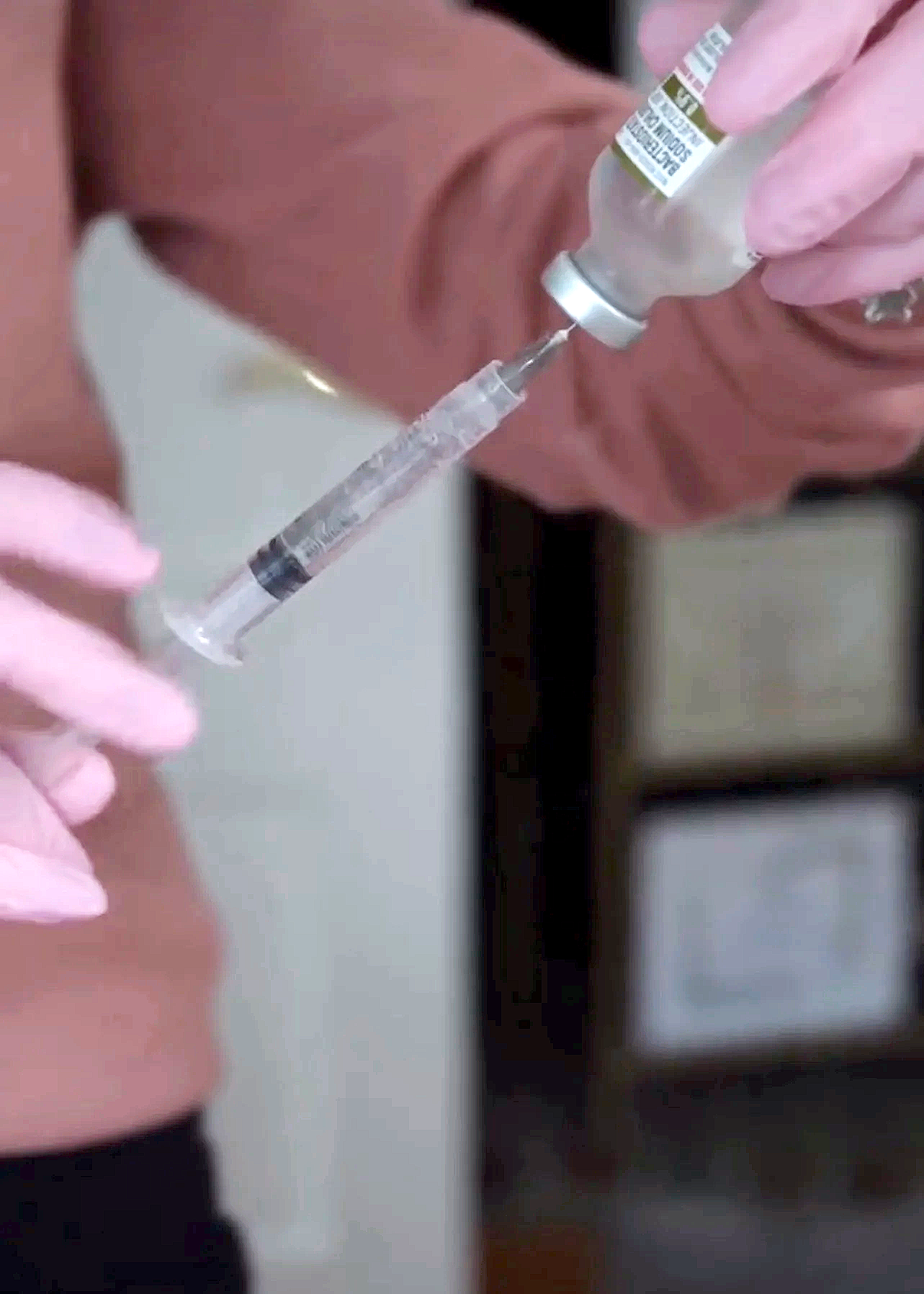 Close-up of a nurse or medical professional drawing a clear liquid from a small glass vial into a syringe, wearing pink gloves and a peach-colored top.