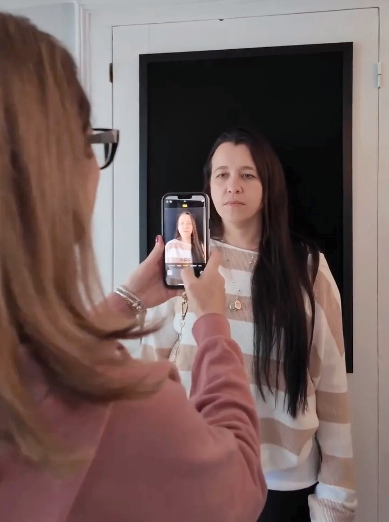 A woman with long dark hair stands in front of a black framed mirror, posing for a photo taken by another woman with reddish hair, glasses, and a pink jacket, using a smartphone.