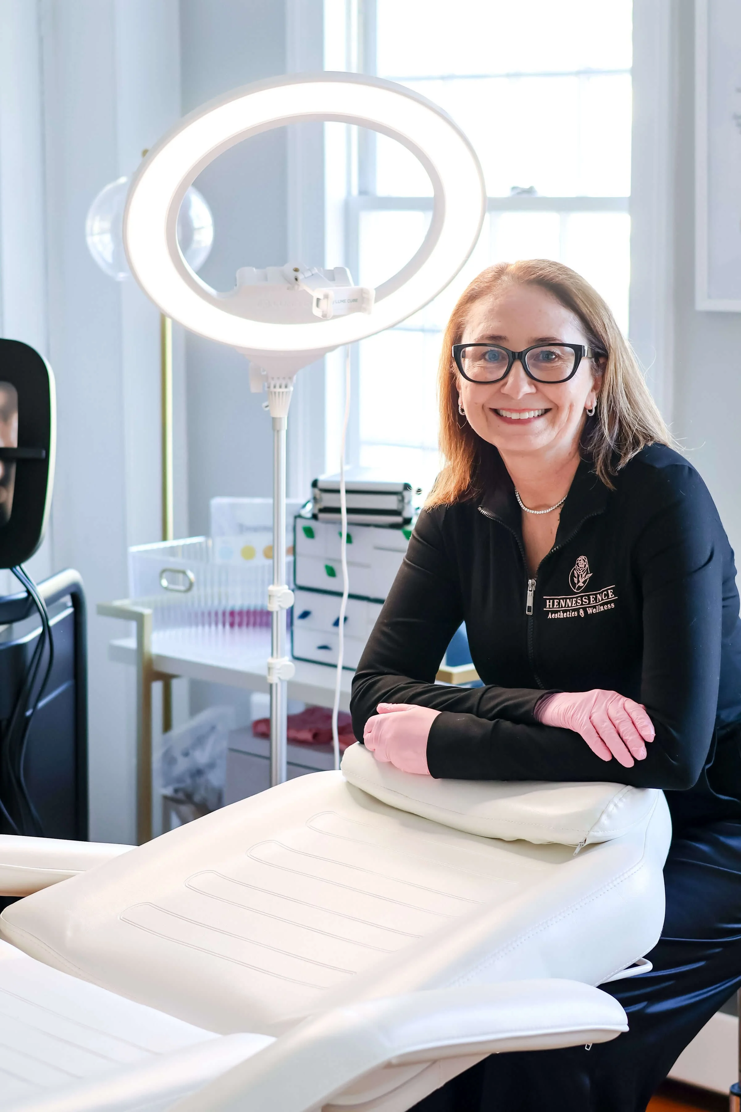 A smiling woman wearing glasses and pink gloves sitting at a white dental or medical chair in a clinic or wellness center, with equipment and supplies in the background.