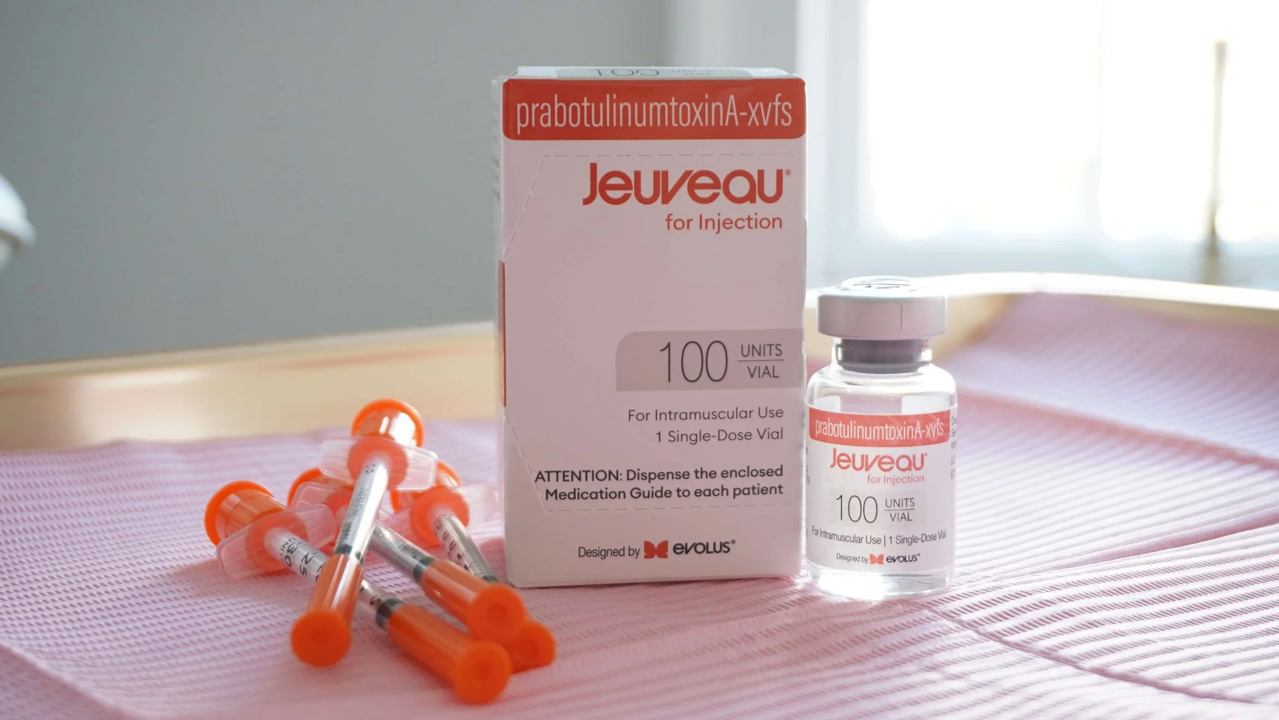 A box and vial of Jeuveau, a brand of botulinum toxin, with several syringes on a pink surface.