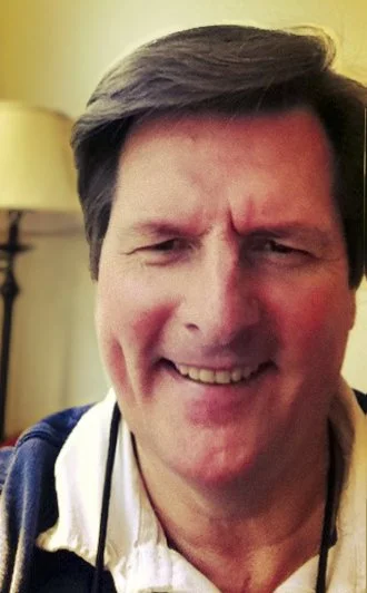 A smiling man with short brown hair, wearing a white shirt and a dark jacket, in a room with a lamp in the background.