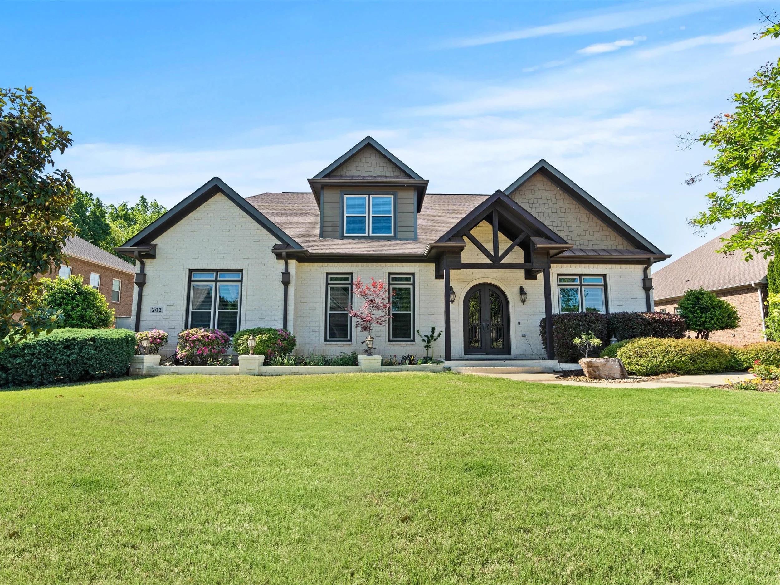 Front view of a large two-story house with a well-maintained lawn, flowering shrubs, and trees, under a clear blue sky.